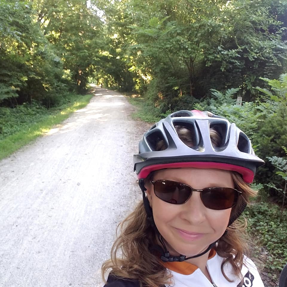 Person wearing a helmet and sunglasses outdoors on a forest path, smiling, with trees and greenery around.