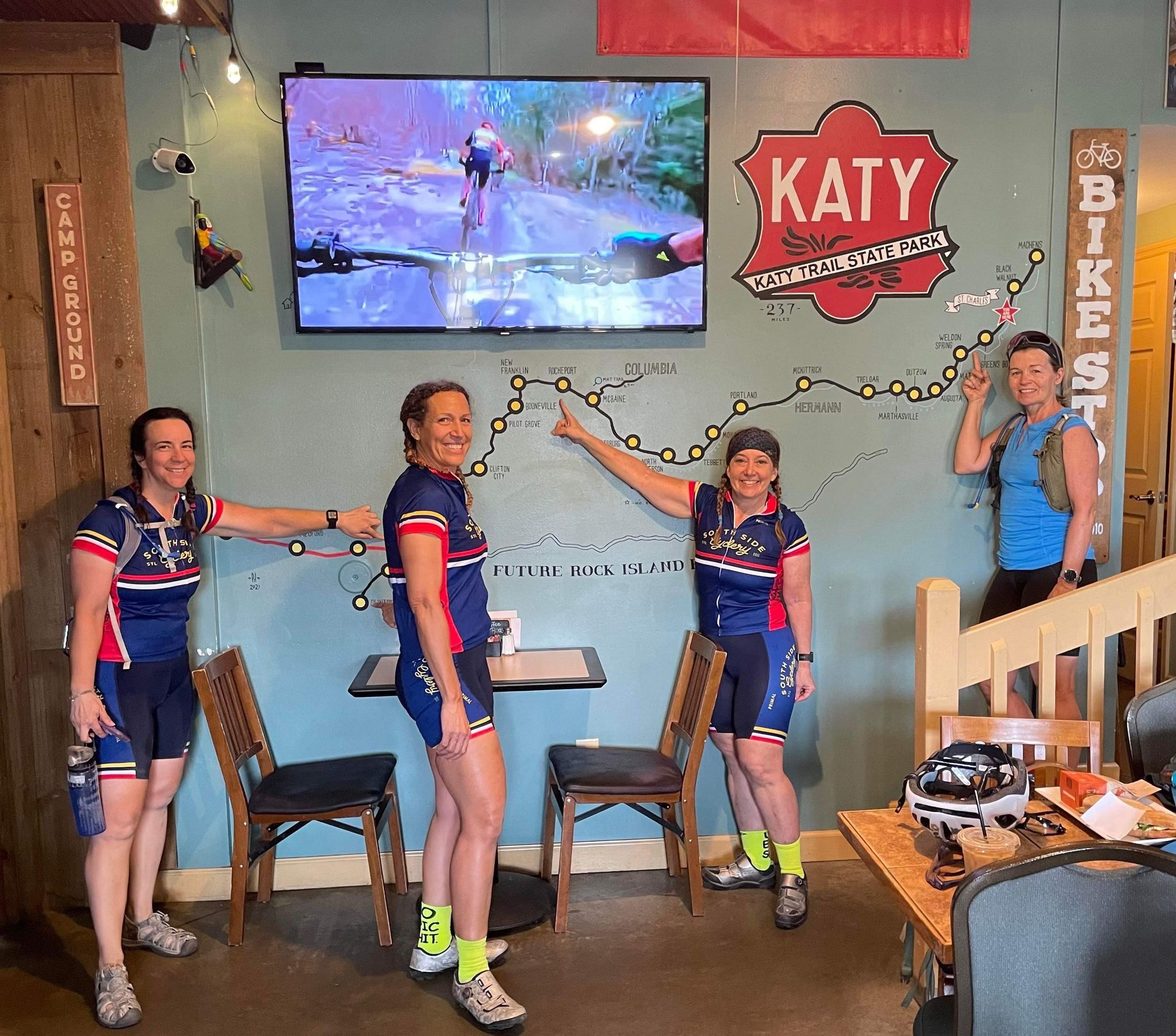Four women in cycling uniforms stand inside a room with a wall decorated with bike-themed art and a TV screen, smiling.