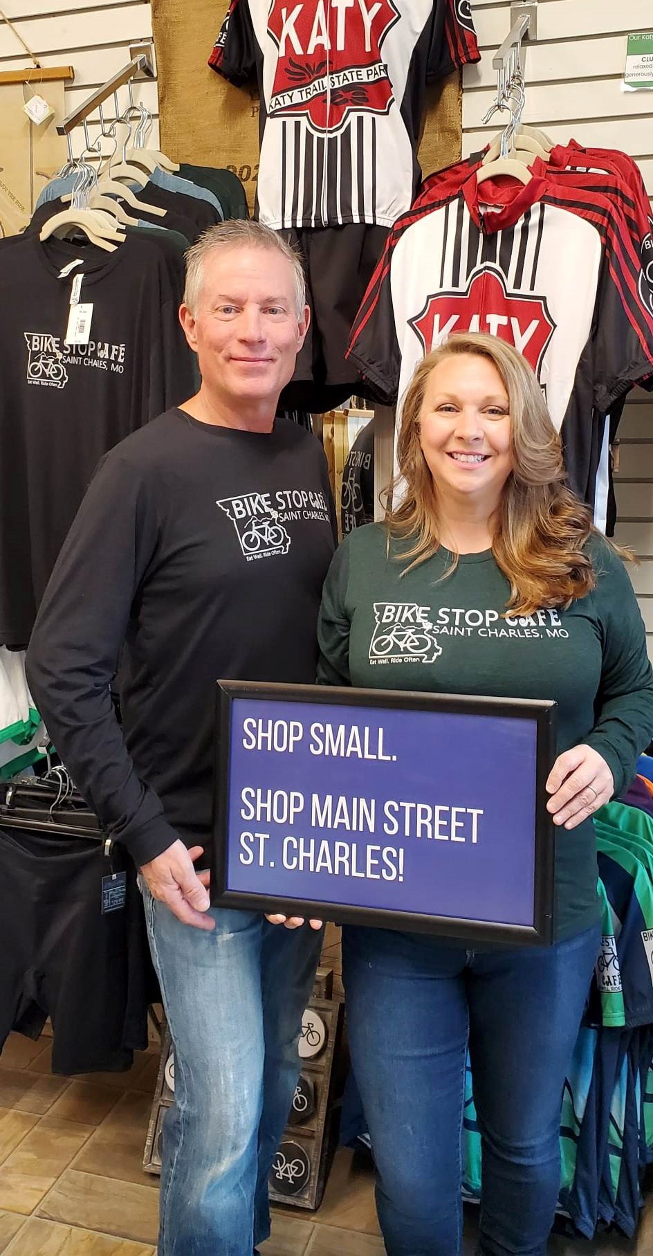 Two smiling people holding a sign in a store with sports jerseys and clothing displayed behind them.