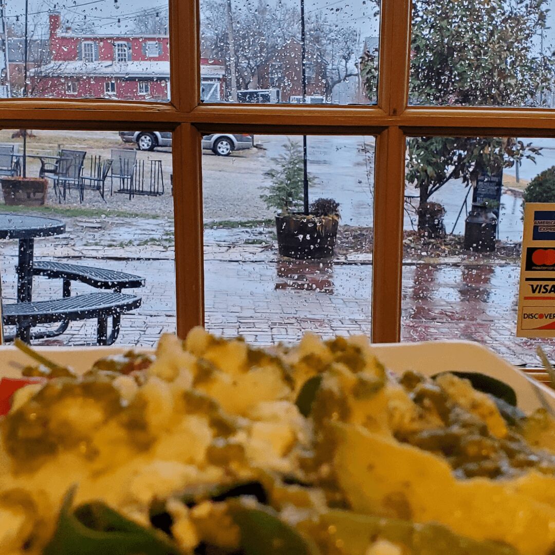 View through a window showing a rainy outdoor scene with wet pavement, trees, and parked cars, with a blurry foreground of food.