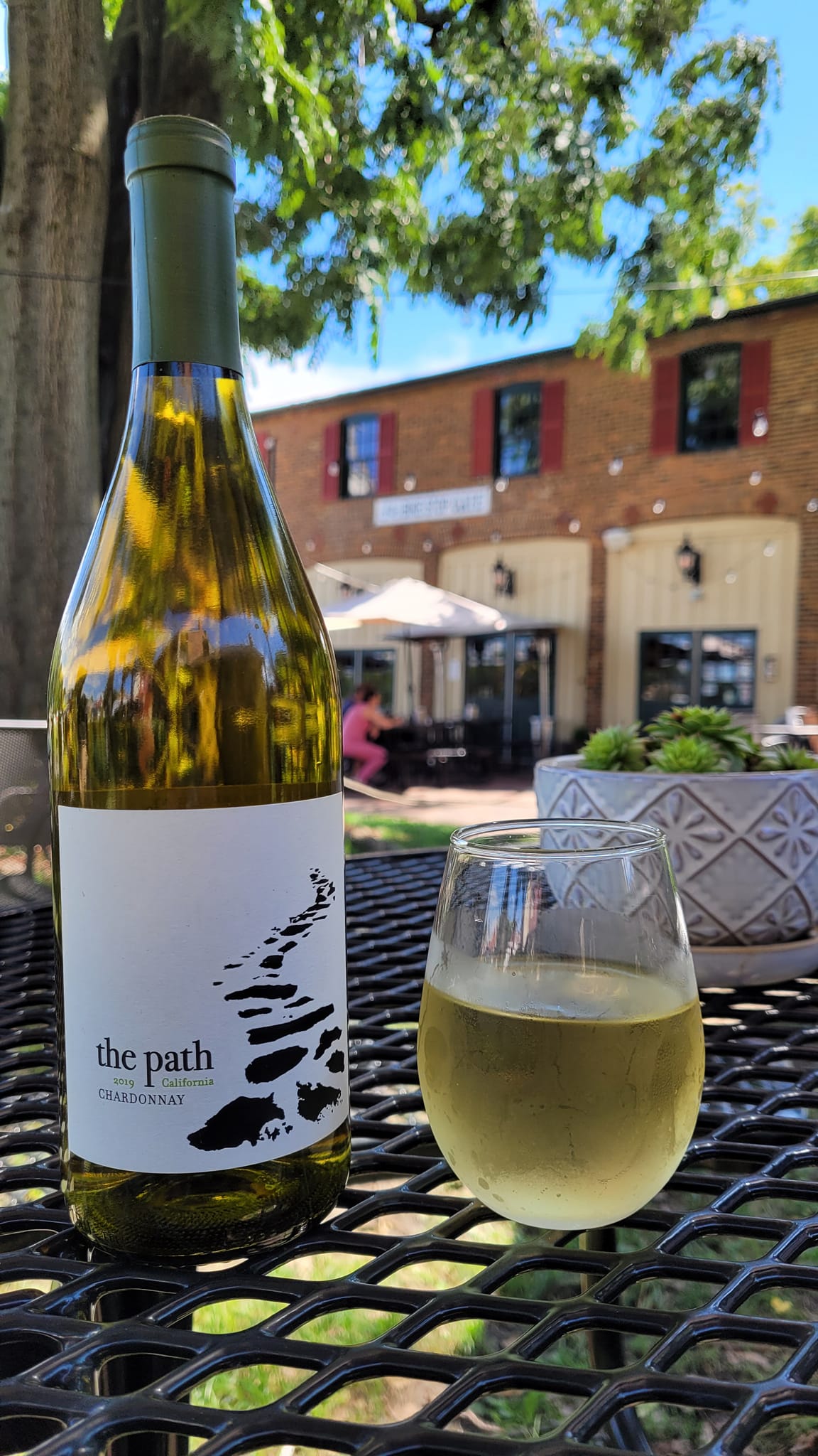 A bottle of white wine and a glass of wine on a black metal table outdoors, with a building and trees in the background.