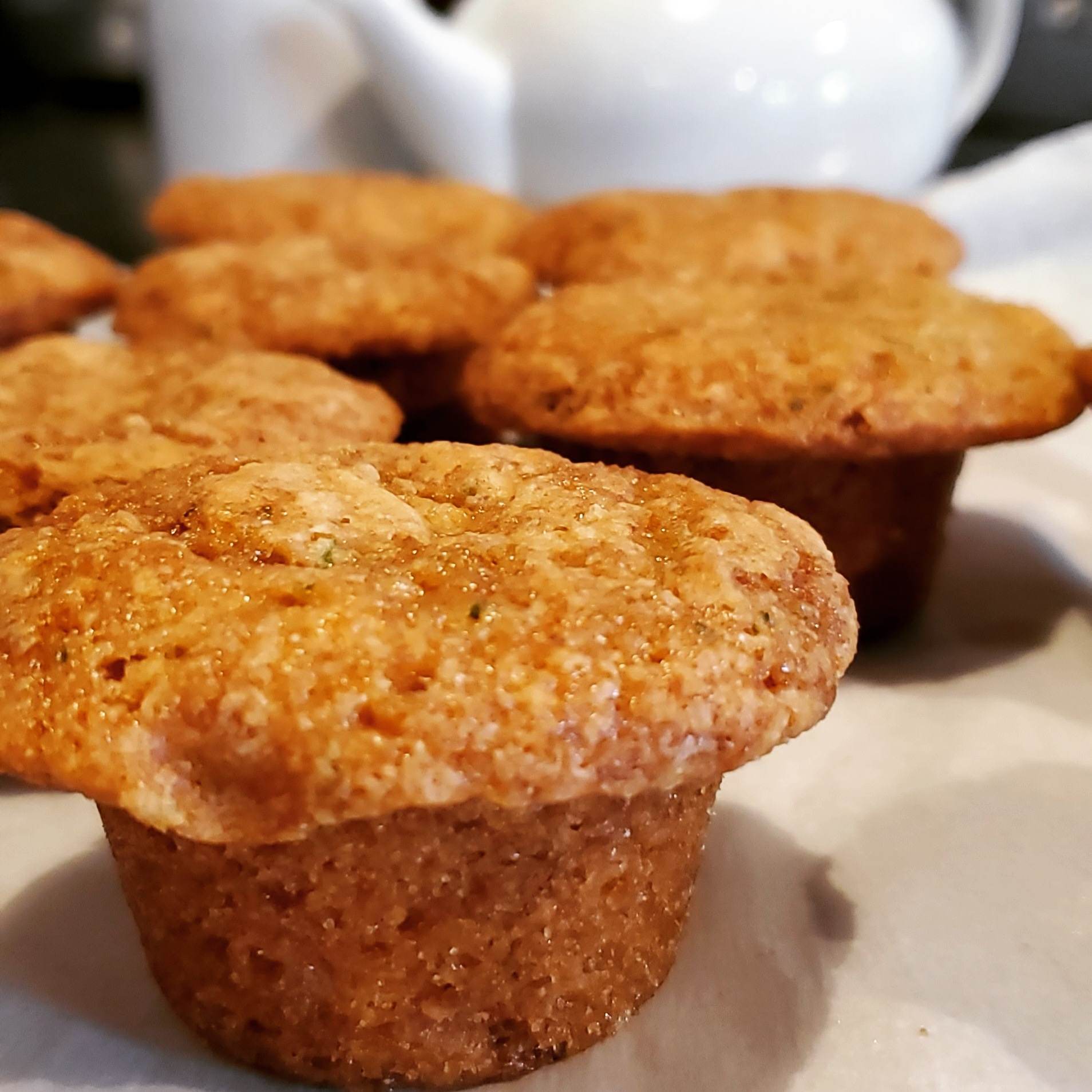 Close-up of golden-brown muffins on parchment paper with a white teapot in the background.