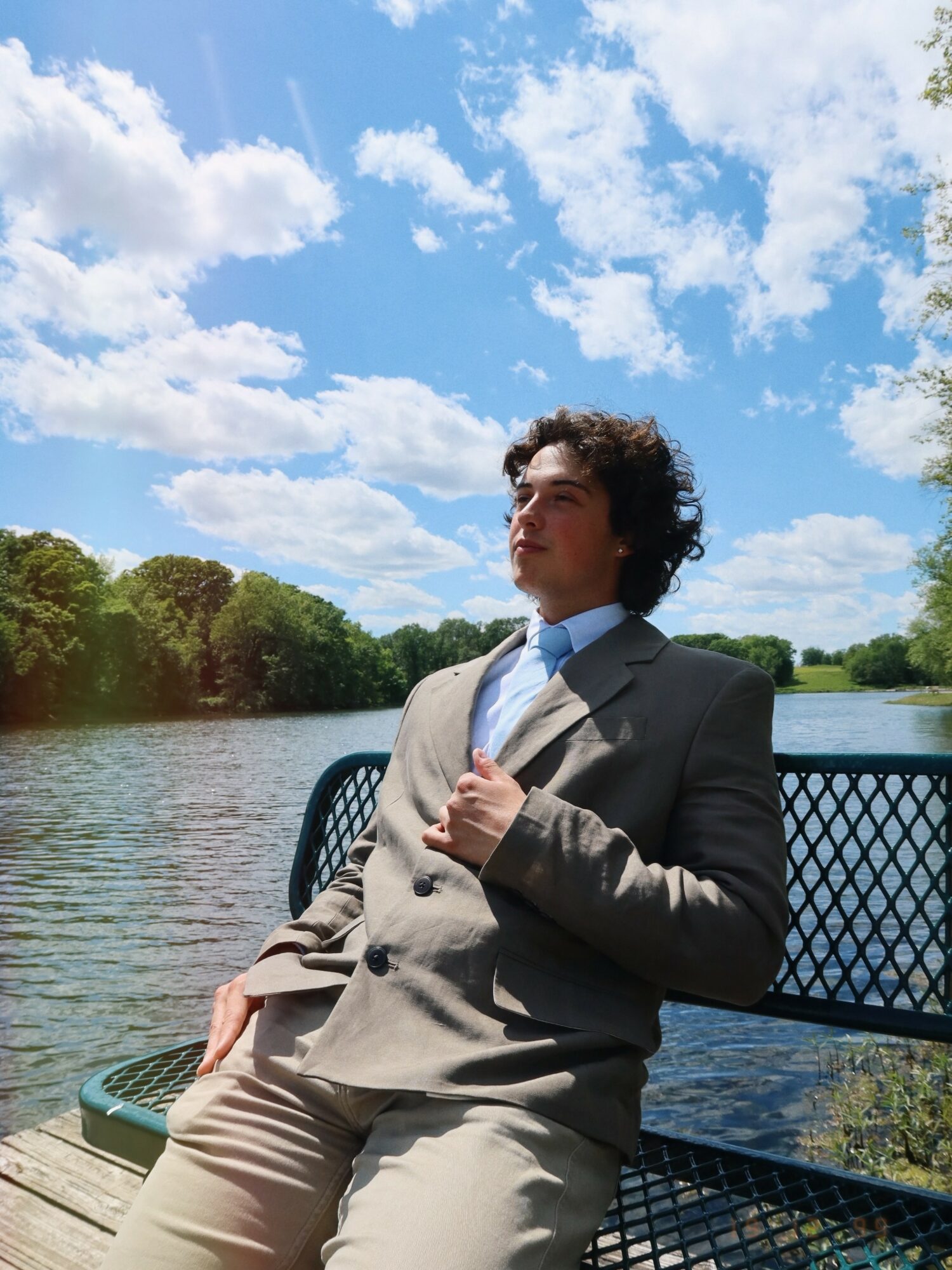Young man in a suit sitting on a bench by a river under a blue sky with clouds, trees in the background.