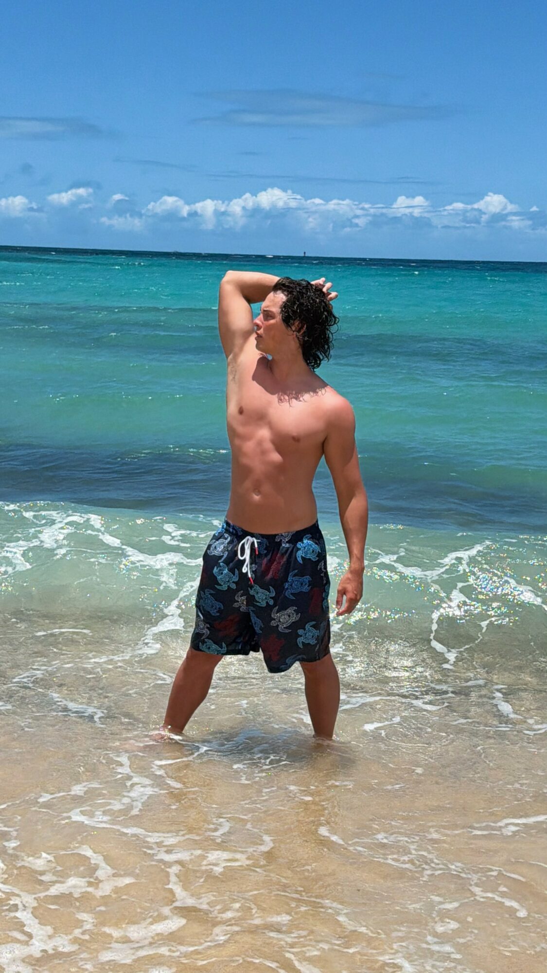 Person standing in shallow ocean water with hand on head, wearing floral swim shorts, facing the sea under a blue sky.