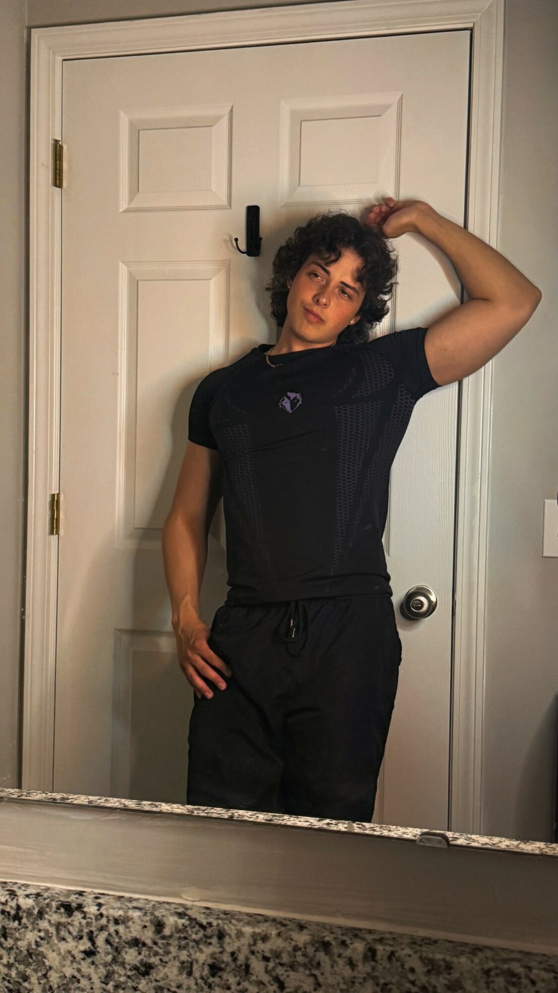 Person with curly hair wearing a black t-shirt and pants, standing in front of a white door, hand on head.