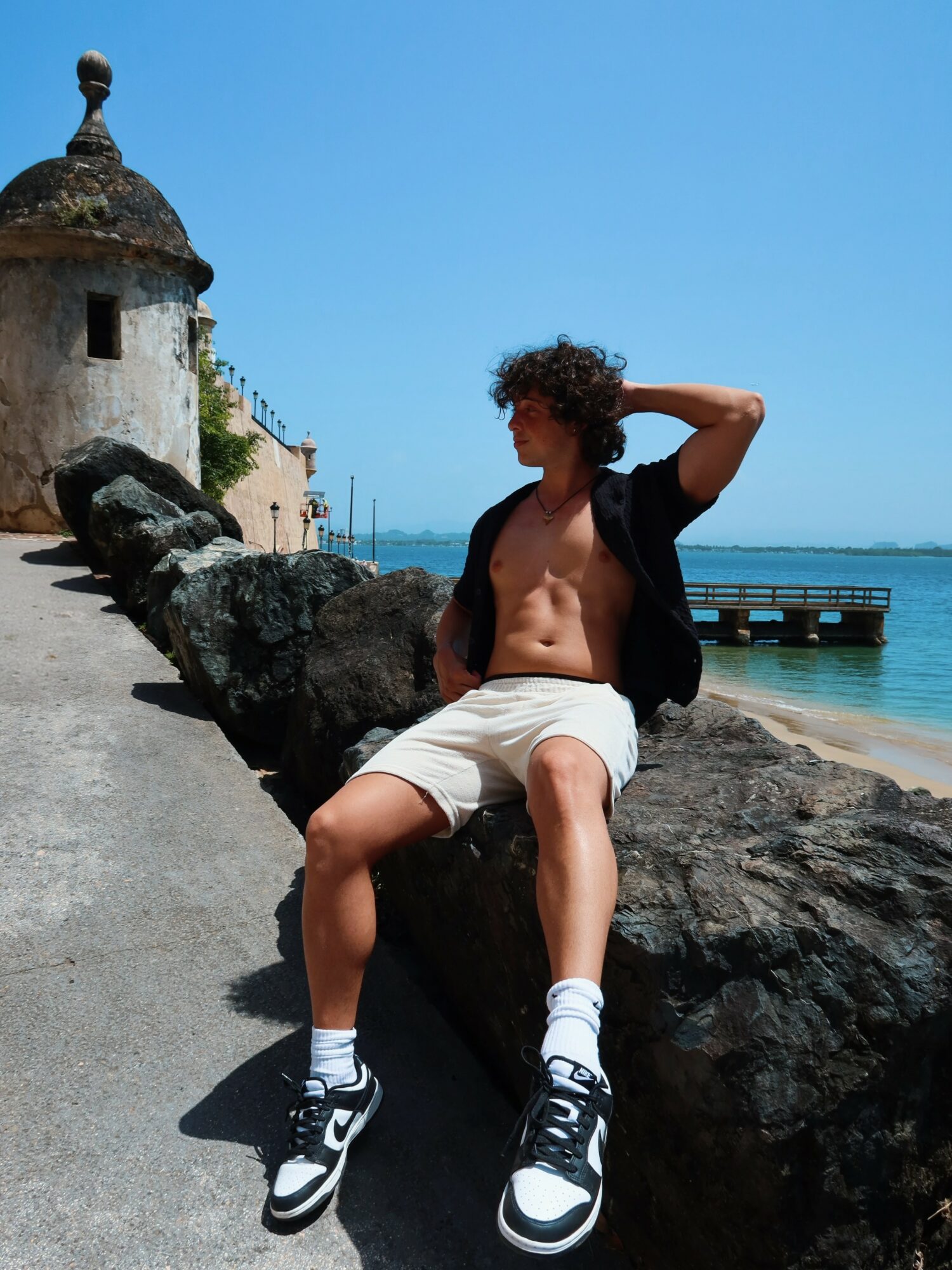 Young man with curly hair sitting on a large rock near the beach, wearing shorts, sneakers, and a black shirt open at the front, with a historic building and pier in the background.