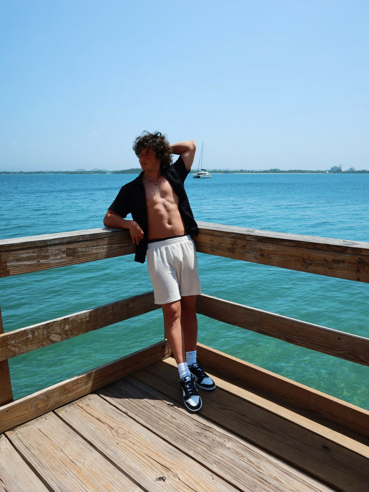 Young person with curly hair leaning on a wooden railing by the water, shirtless, wearing white shorts and sneakers, under a clear blue sky.