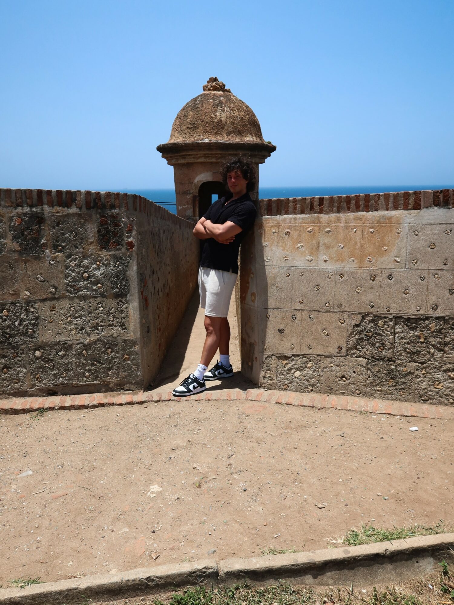 Person standing with arms crossed in front of a stone fortress wall and tower, clear blue sky above.