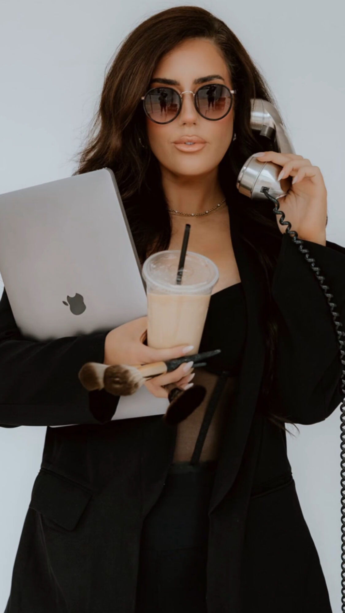 Woman with sunglasses holding a phone, laptop, and drink, wearing a black blazer, against a light background.