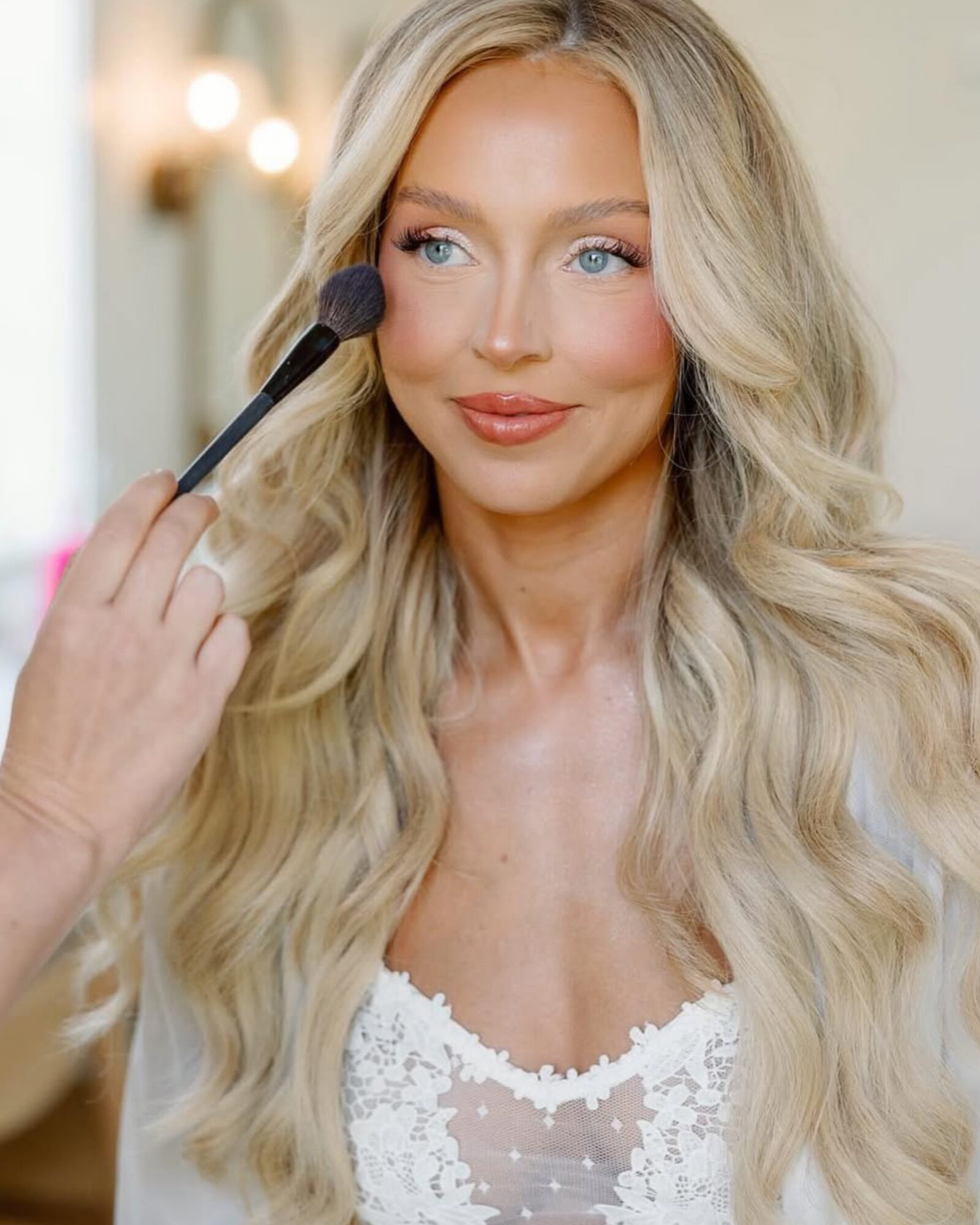 Woman with long blonde hair having makeup applied with a brush, smiling softly, indoors with warm lighting