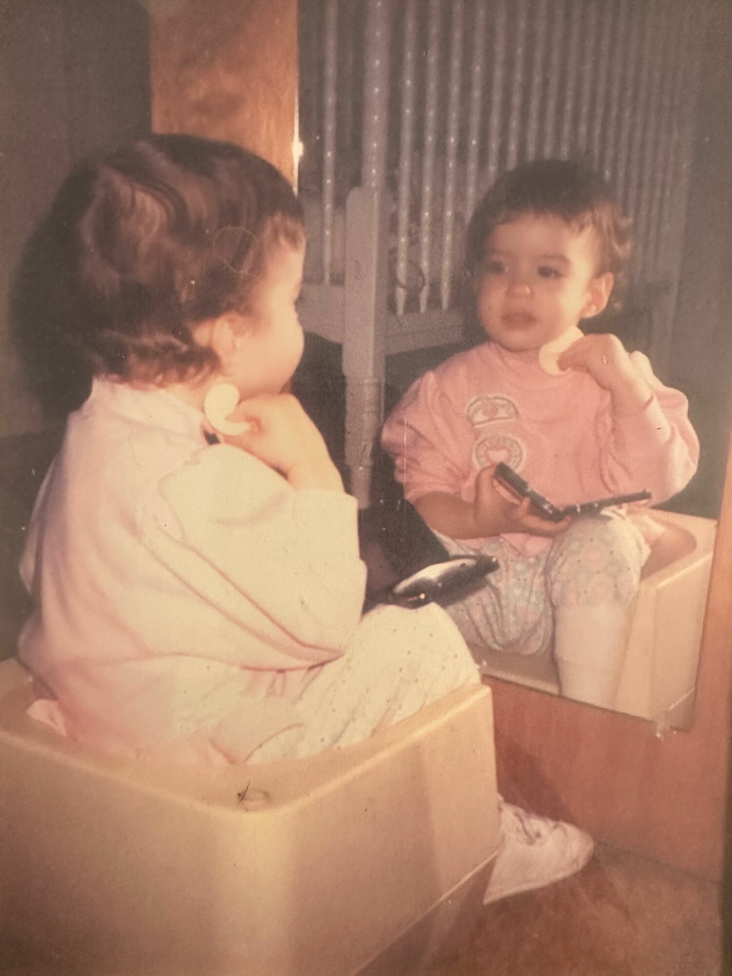 Child sitting on a chair holding a doll, with another person sitting nearby, in a room with curtains and wooden furniture.