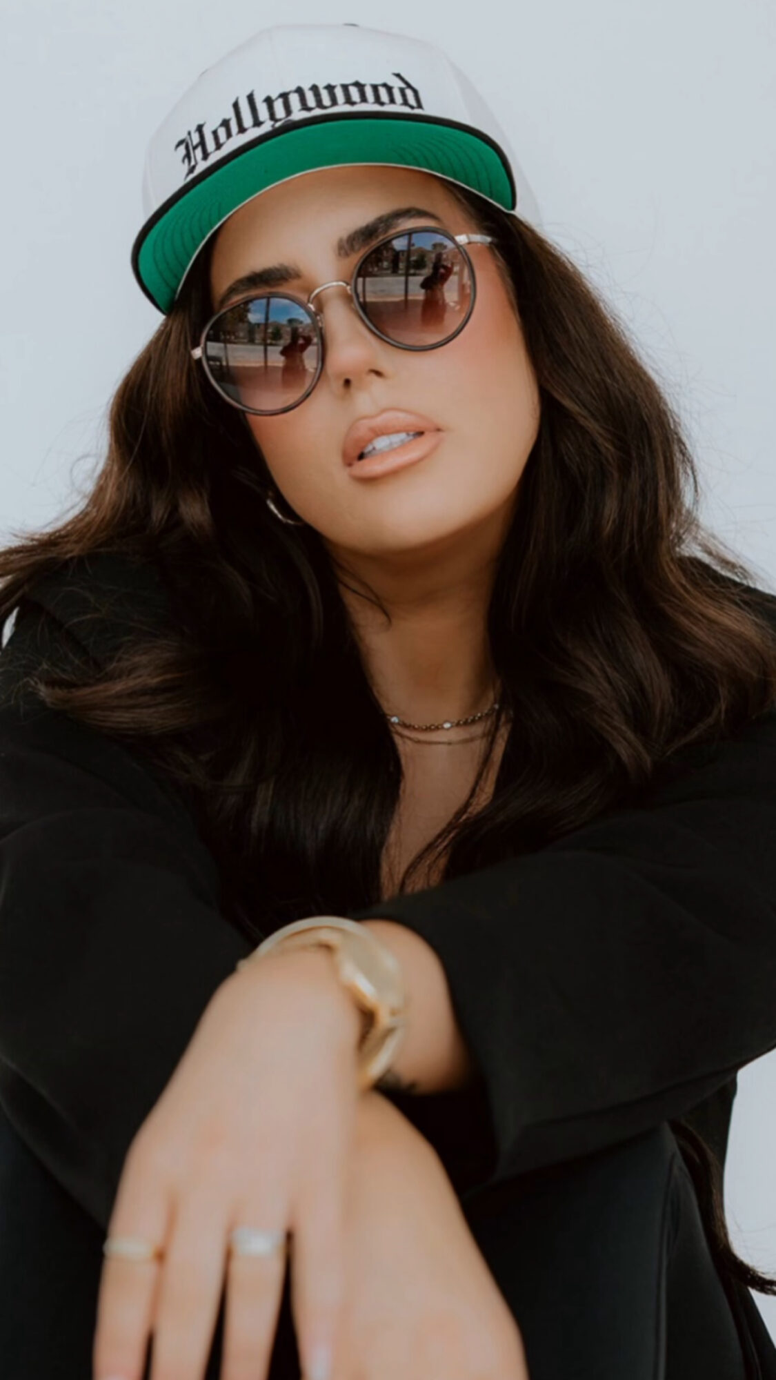 Woman wearing sunglasses, a cap, and a black jacket, with long dark hair and a neutral expression.