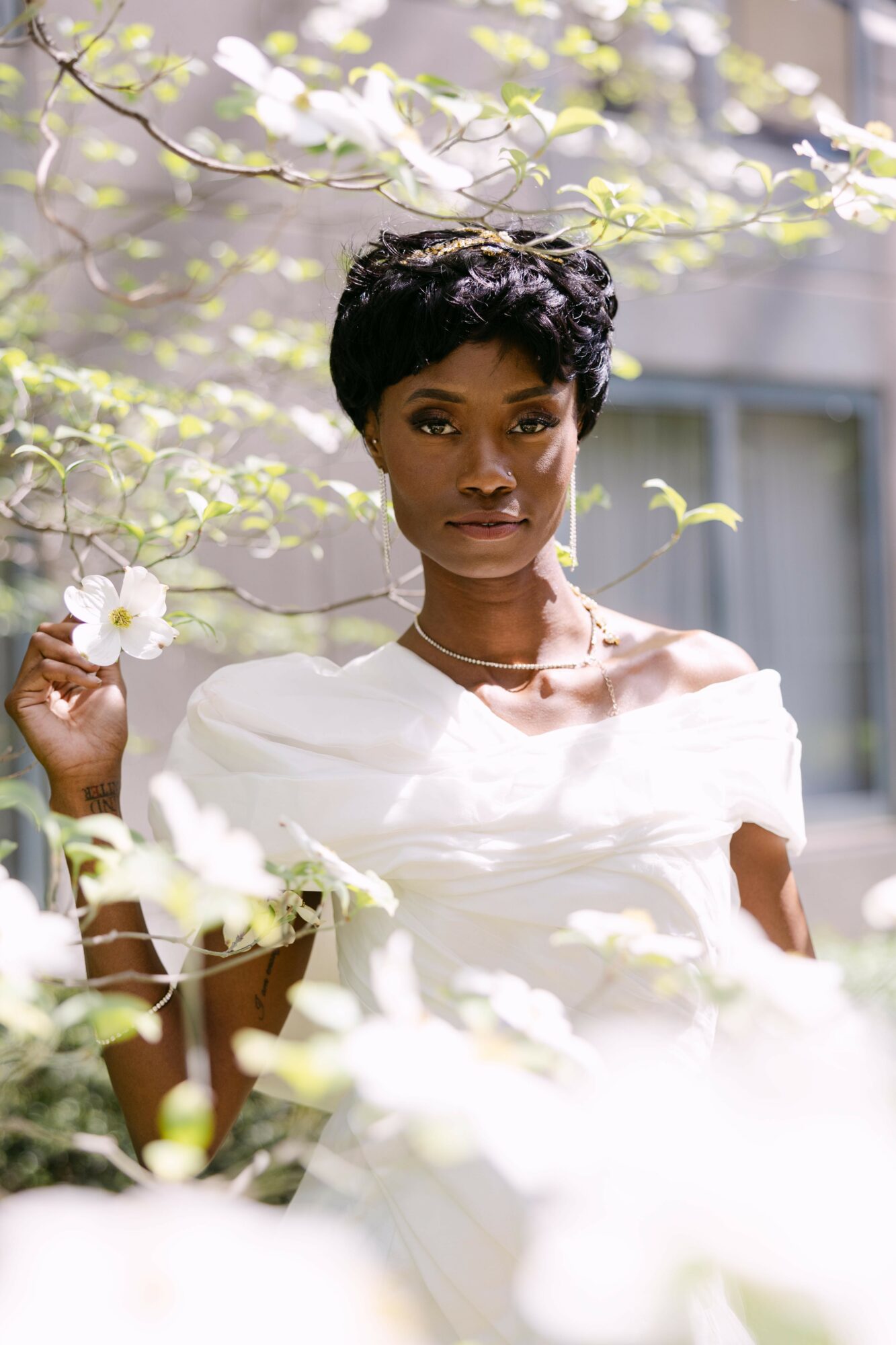 Woman with short curly hair in a white dress outdoors among blooming branches, sunlight, and a building in background.
