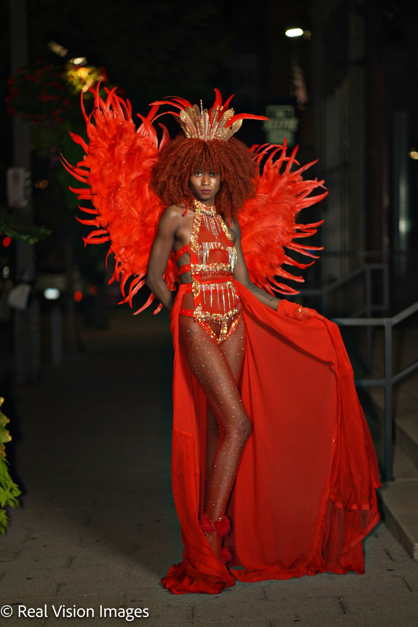 Person in elaborate red costume with large feathered wings and a flowing cape, standing outdoors at night.