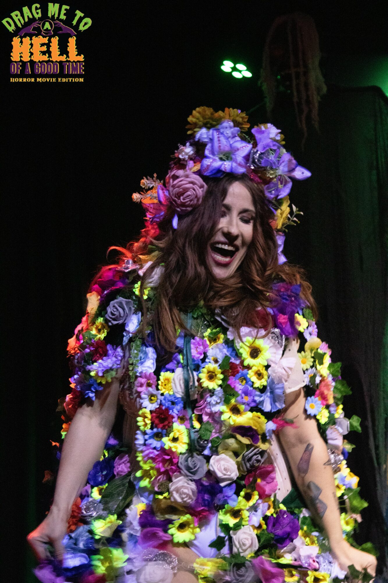 Woman with a large floral headdress and dress made of colorful flowers, smiling on stage with a dark background.