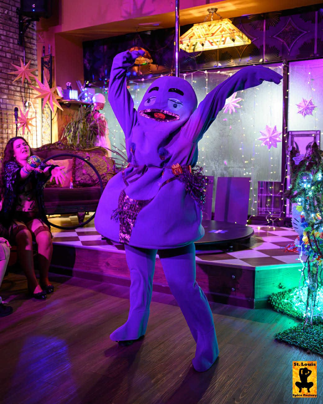 Person in a purple costume dancing with arms raised in a decorated room with Christmas trees and lights.