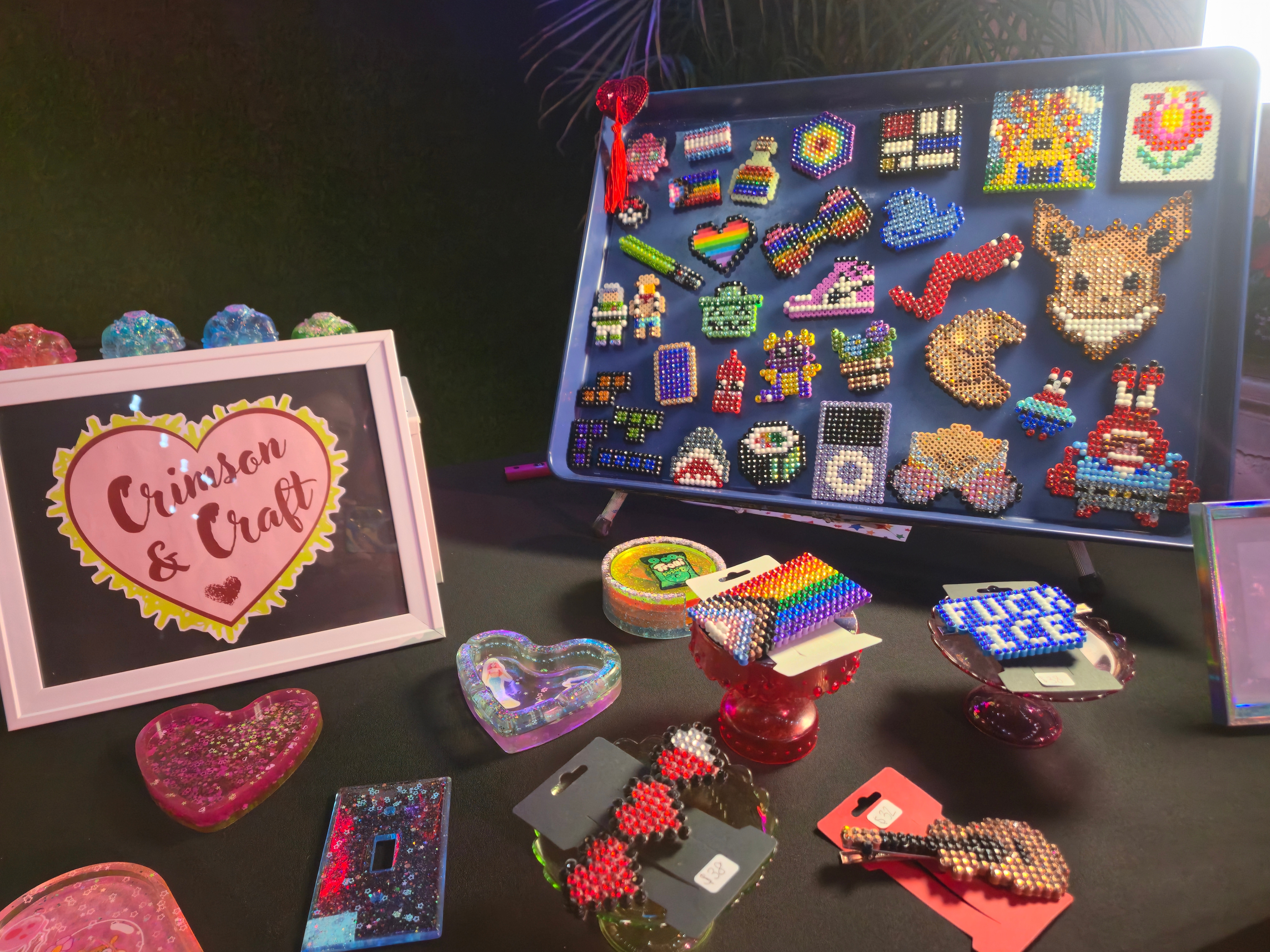 Display of colorful pixel art crafts, a pink sign with heart and text, and various heart-shaped items on a black surface.