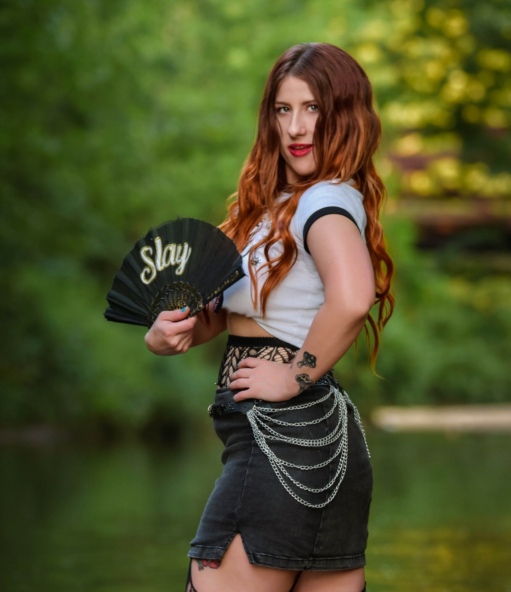 Young woman with long red hair holding a black fan with 'Stay' written on it, standing outdoors near water.