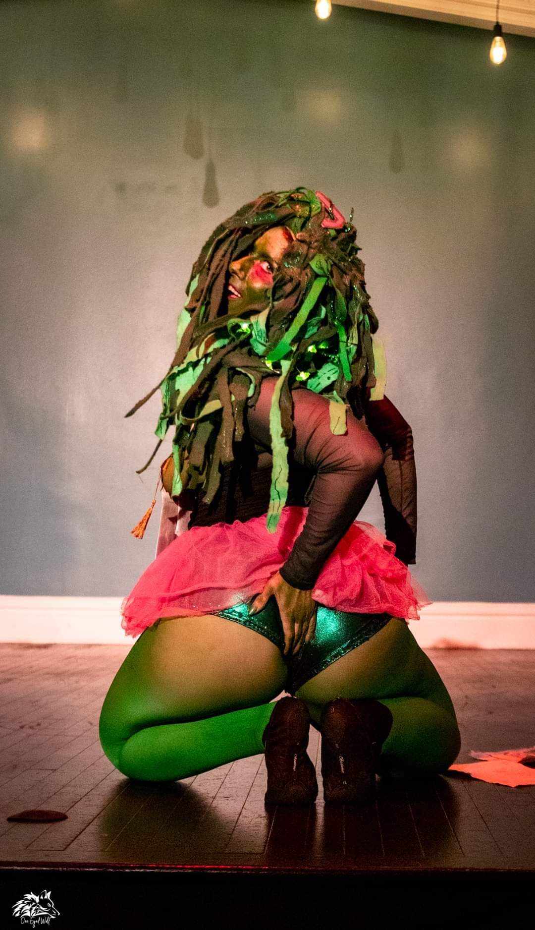 Person with colorful dreadlocks crouching on stage, wearing a red skirt, black top, and green tights, with a plain background.