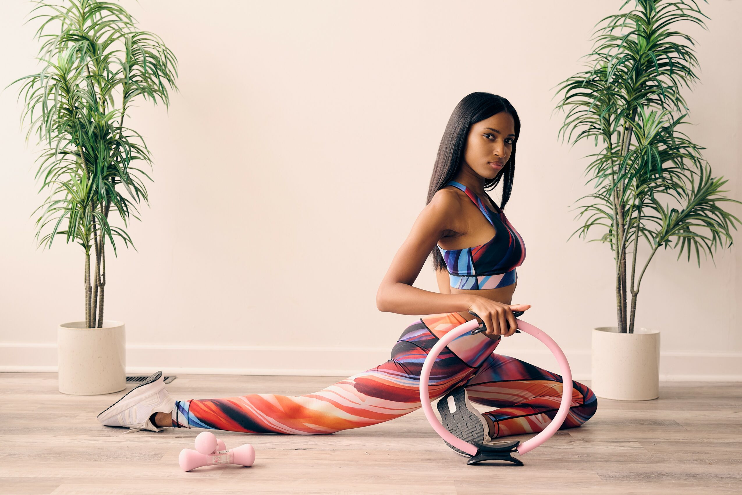 Woman in colorful striped dress kneeling on floor with pink hula hoop, two potted plants behind her, neutral background.