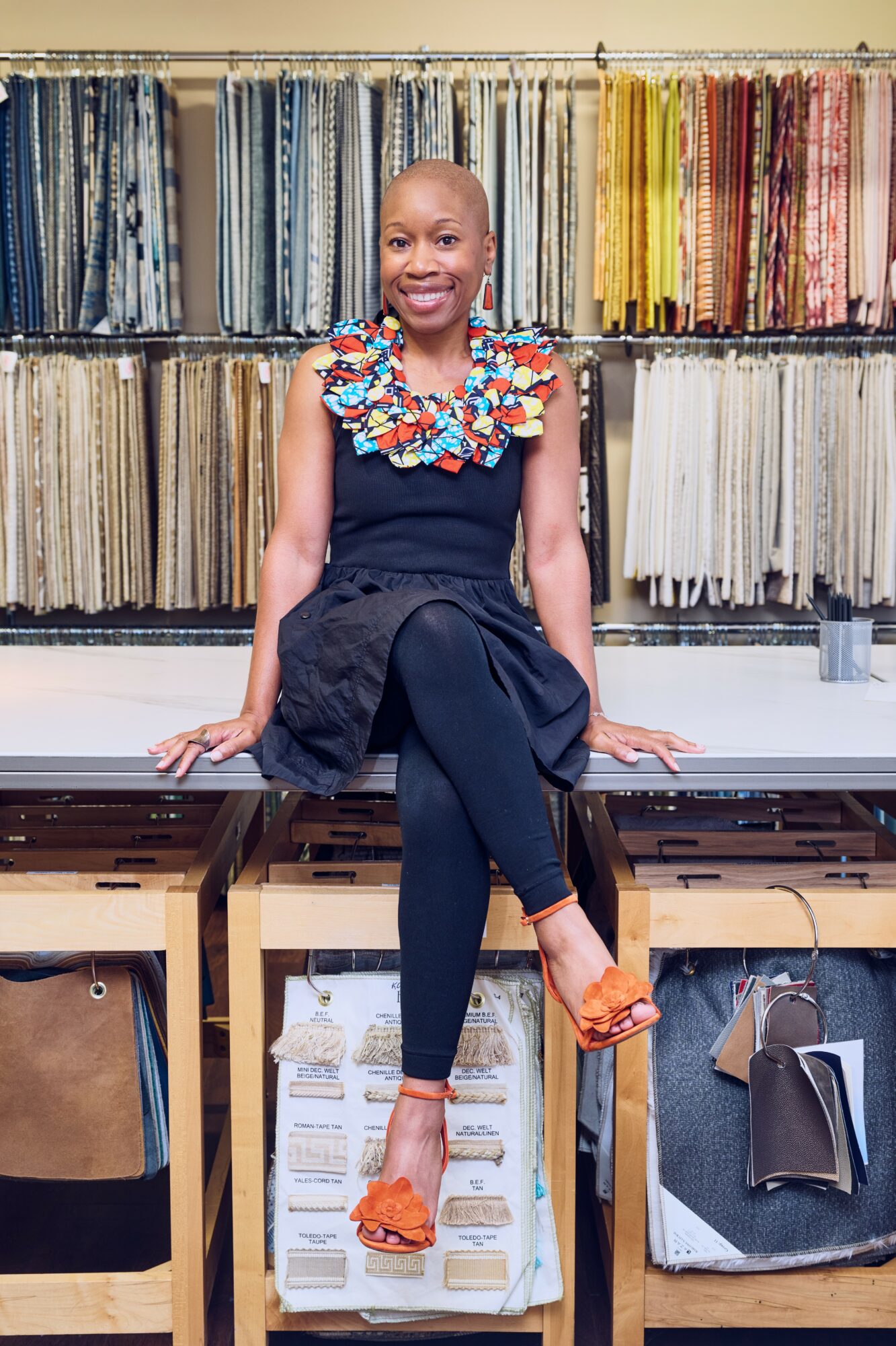 Woman sitting on a table in fabric store, surrounded by colorful fabric samples, smiling, wearing a black outfit with ruffled collar, orange shoes.