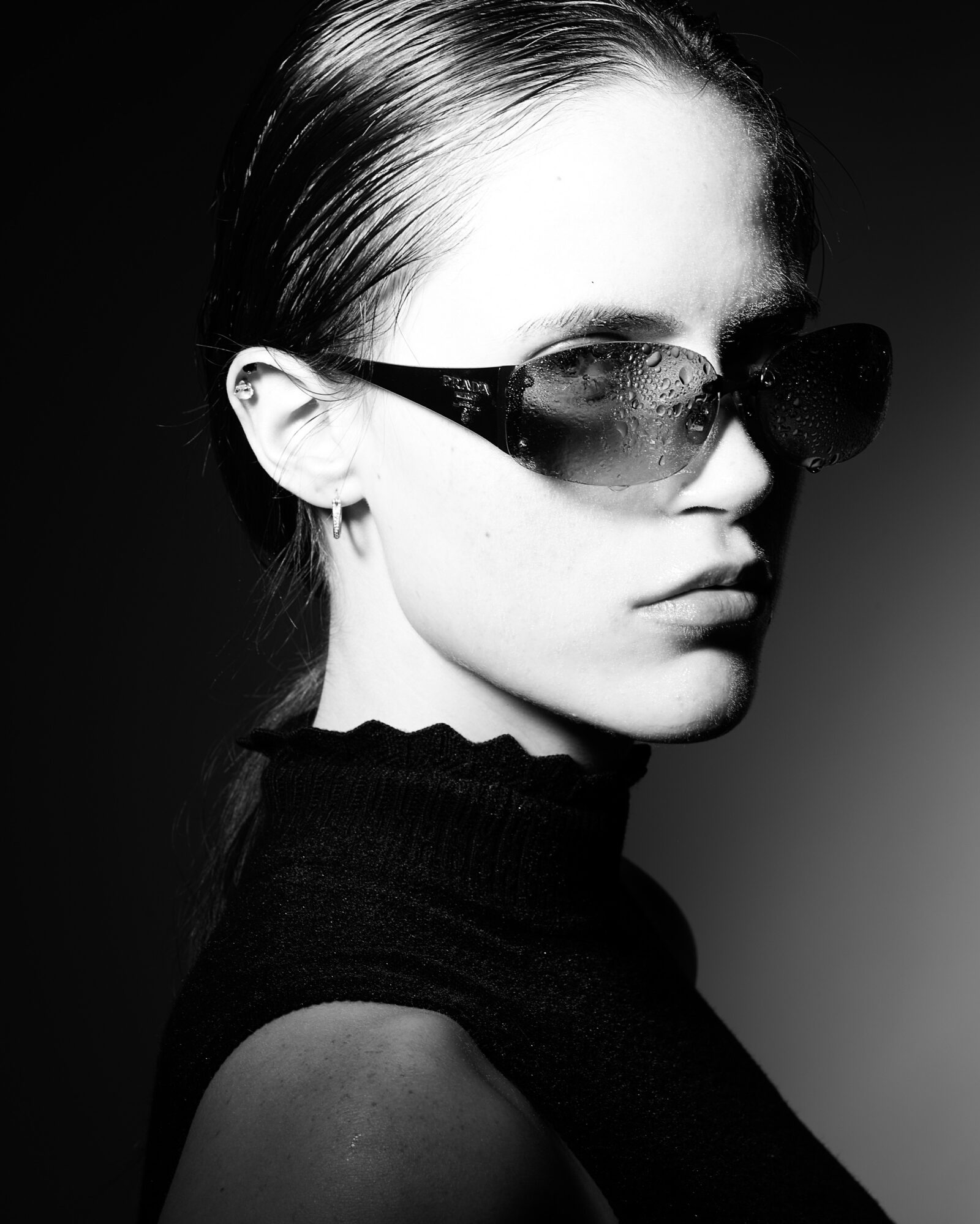 Woman with sunglasses and slicked-back hair, wearing a high-neck top, looking sideways in black and white.