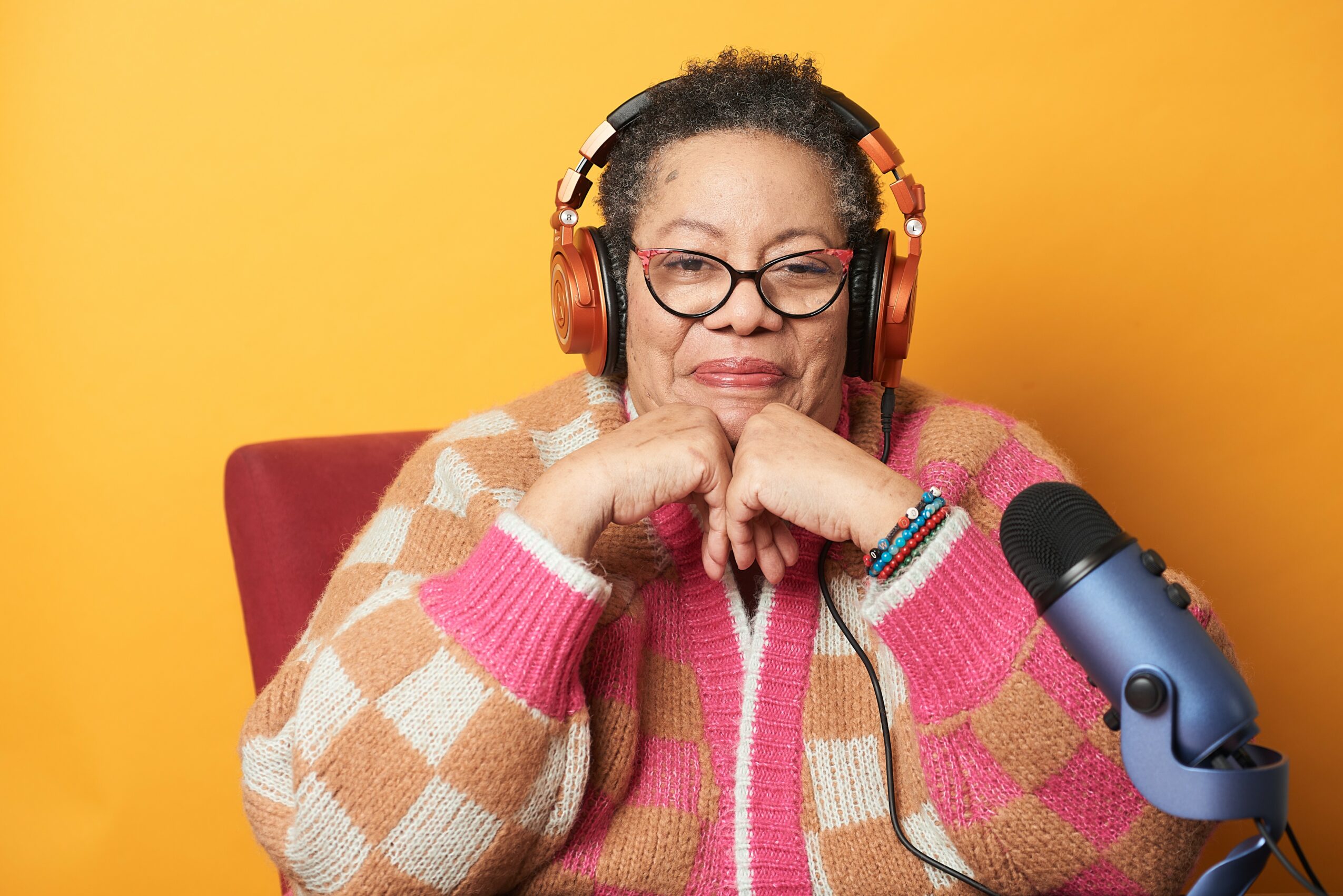 Woman wearing headphones and glasses, smiling with hands under chin, sitting in front of a microphone against a yellow background.
