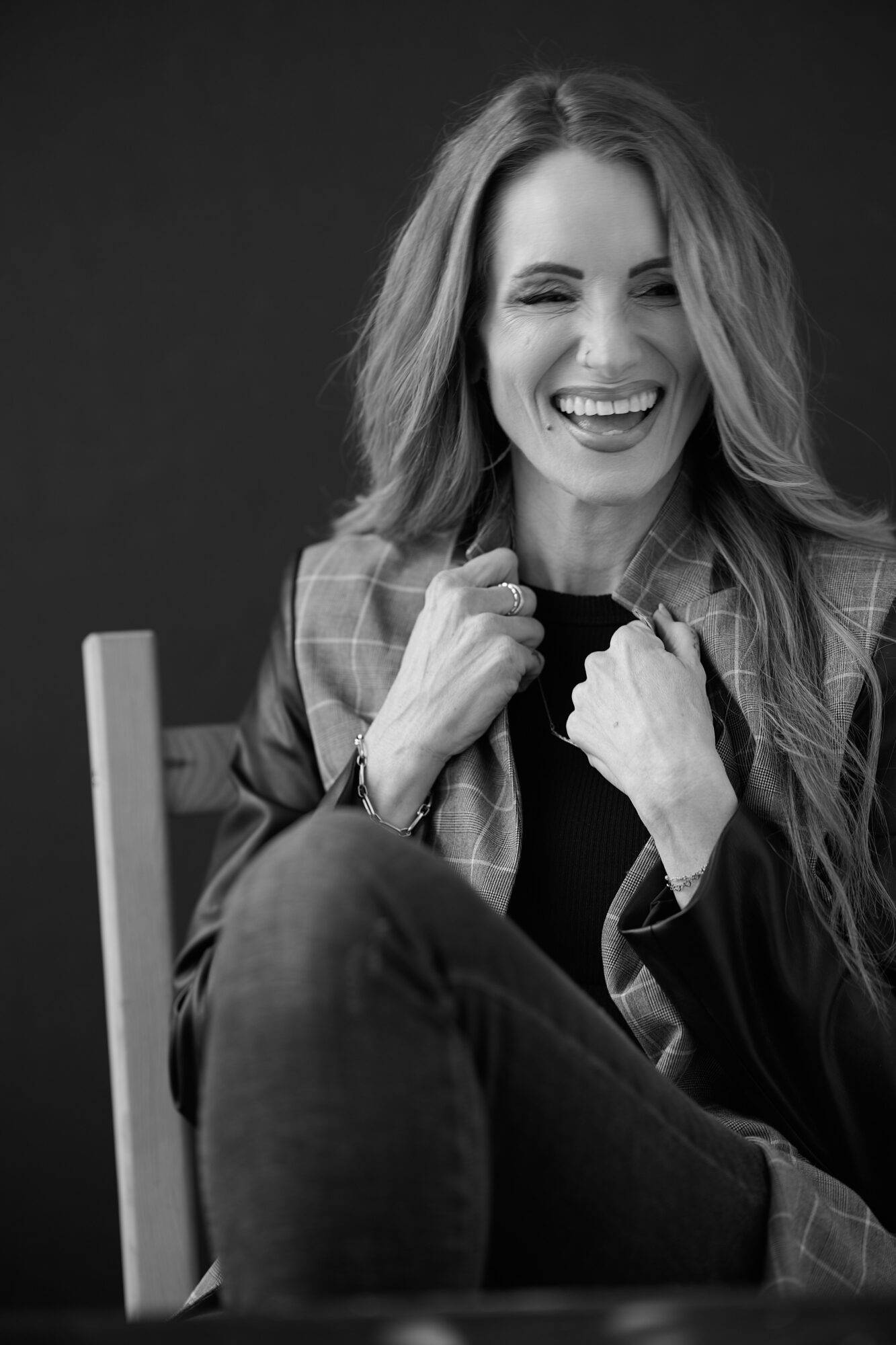 Woman with long hair smiling, sitting with legs crossed, wearing a jacket, in black and white.