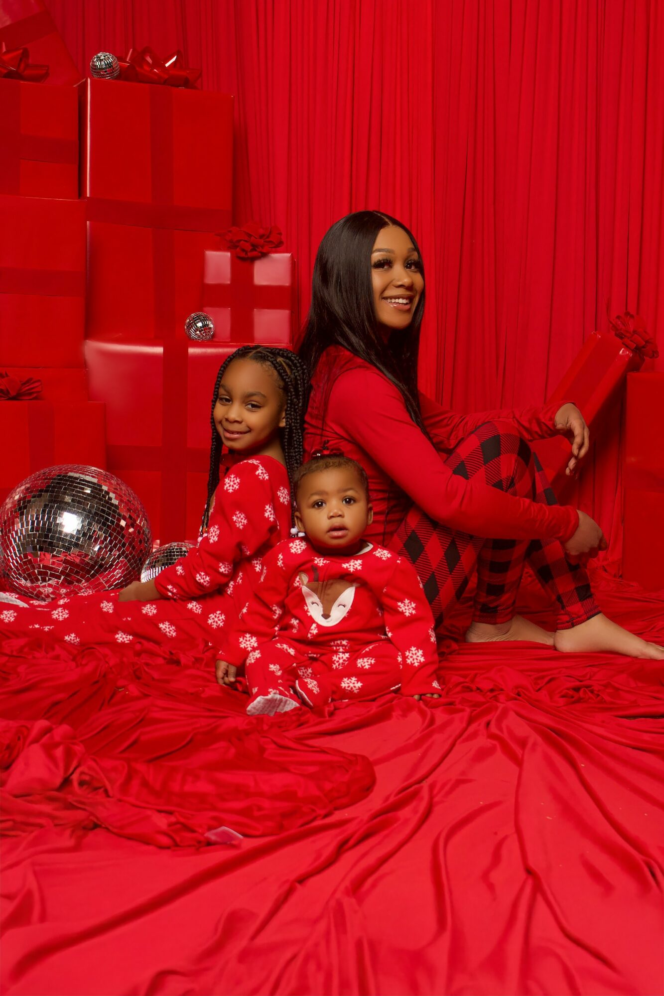 Three people in red pajamas sitting on a red bed with a red curtain background, smiling and posing.