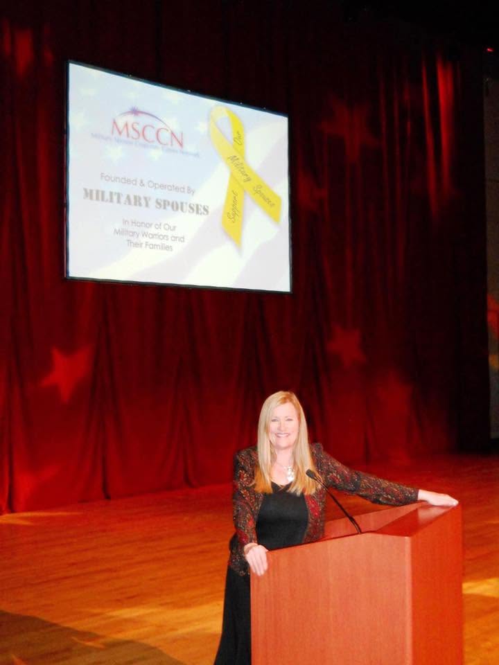Woman standing at podium on stage with red curtains, presentation slide with yellow ribbon behind her.