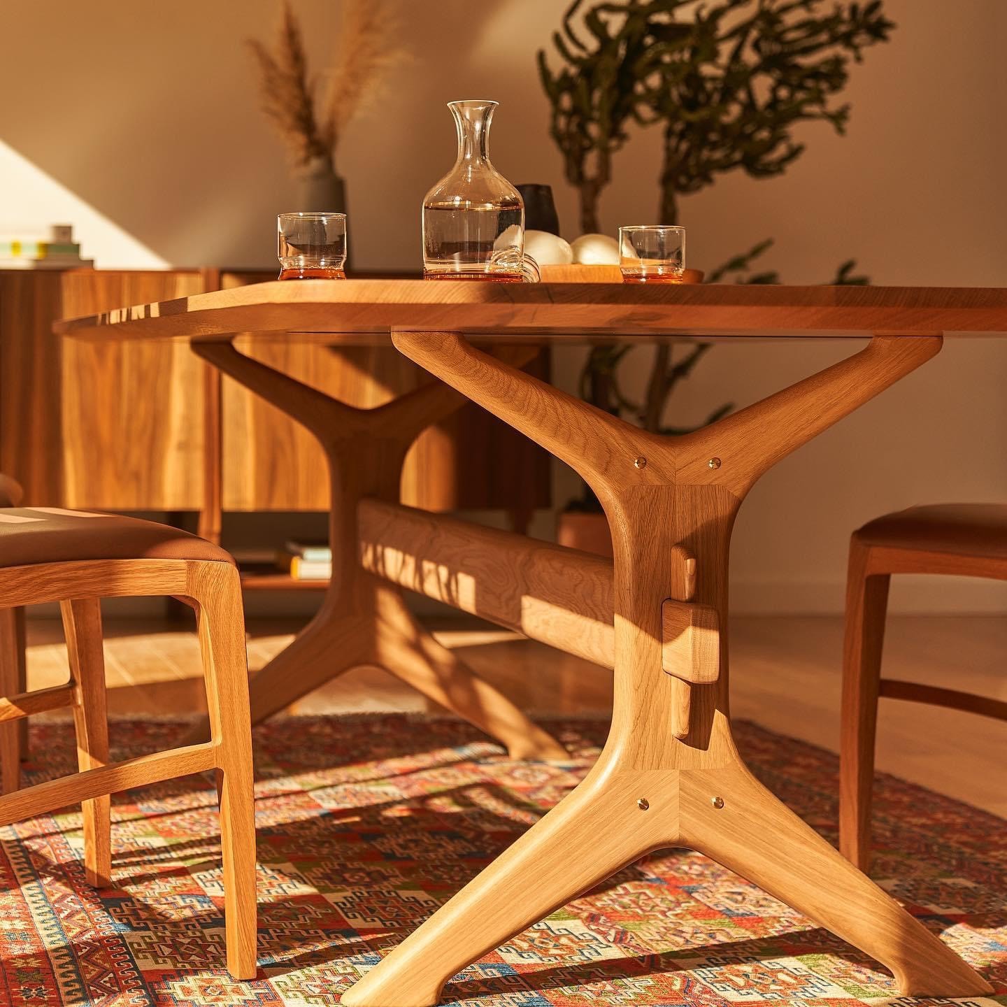 Wooden dining table with chairs, glass pitcher, and glasses, in a room with a patterned rug and plants in the background.