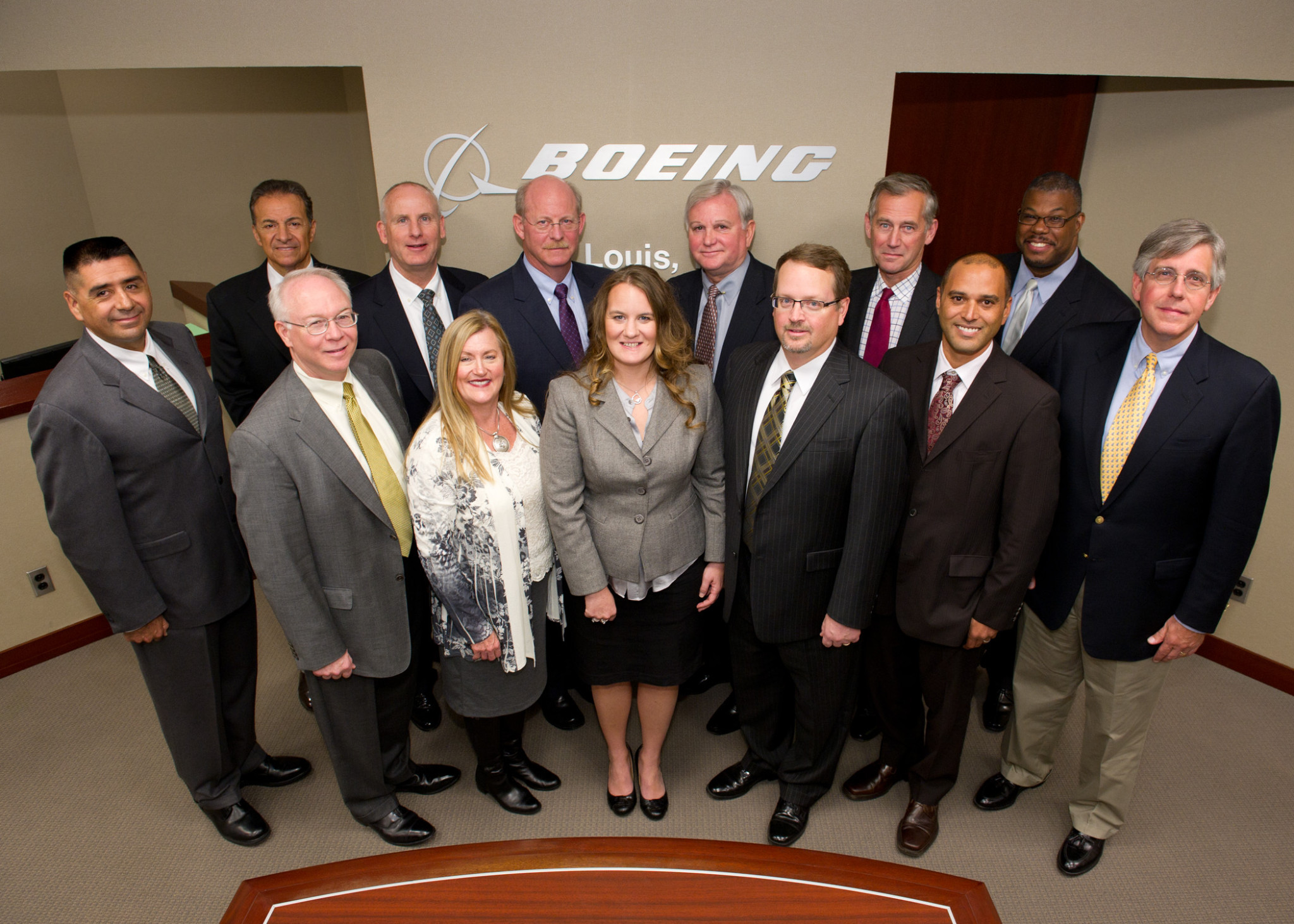 Group of fifteen people standing in front of a Boeing logo, posing for a photo indoors.