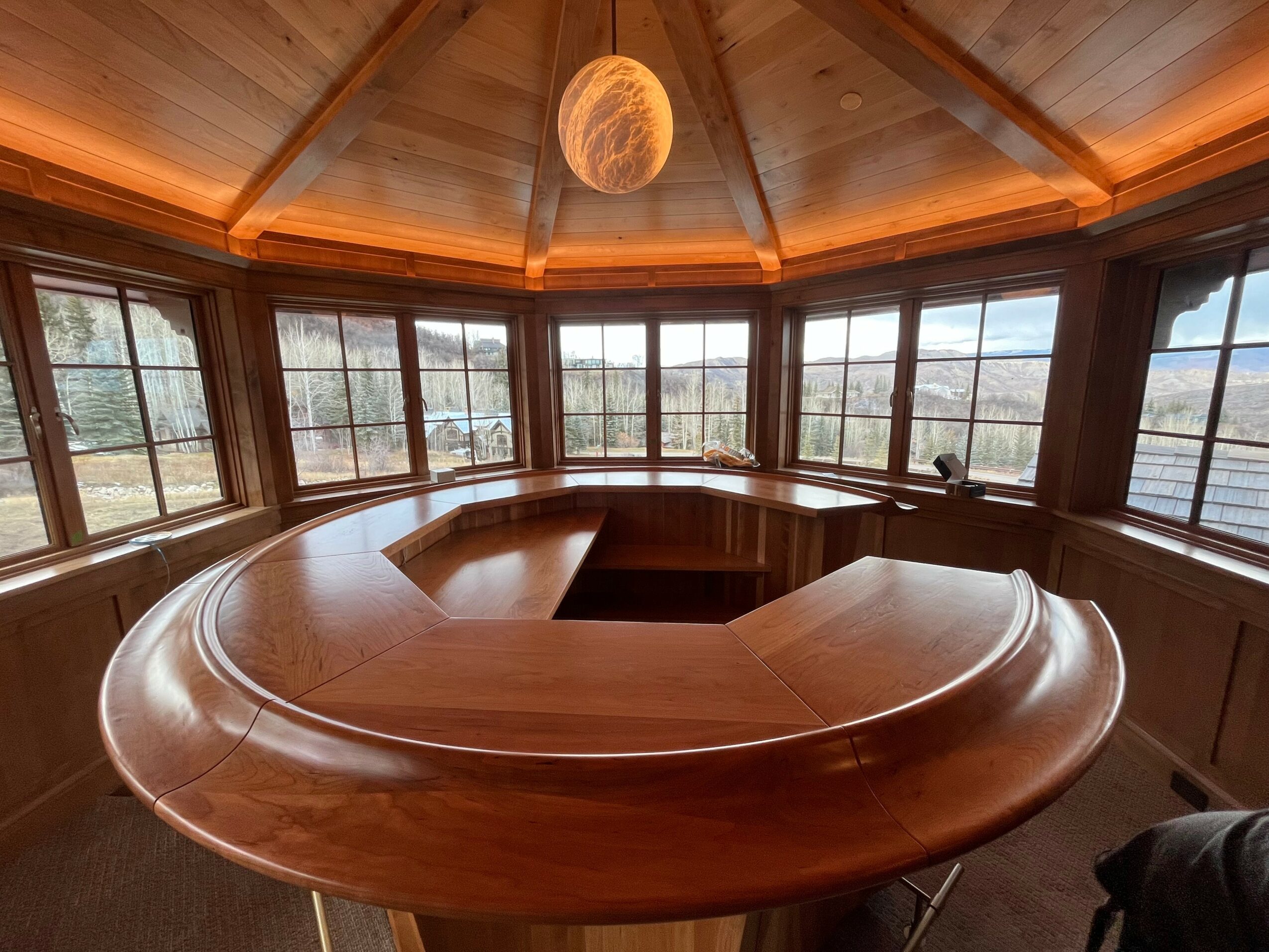 Round wooden room with large windows, a central open space, and a wooden ceiling with a hanging spherical light fixture.