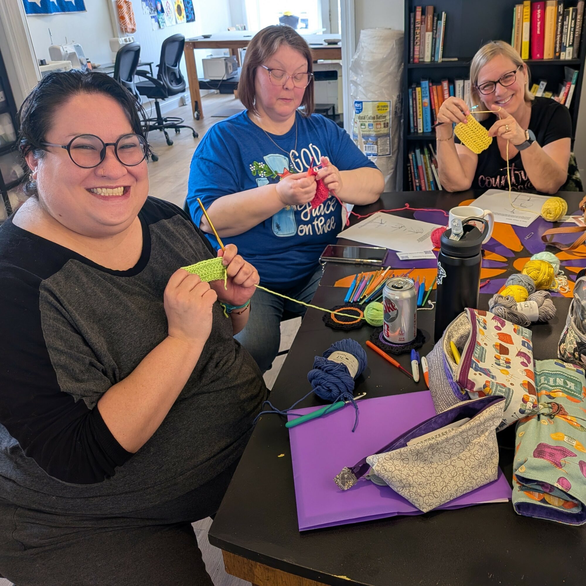 Three women sitting at a table knitting and crafting with yarn, scissors, and supplies, smiling and looking at the camera.