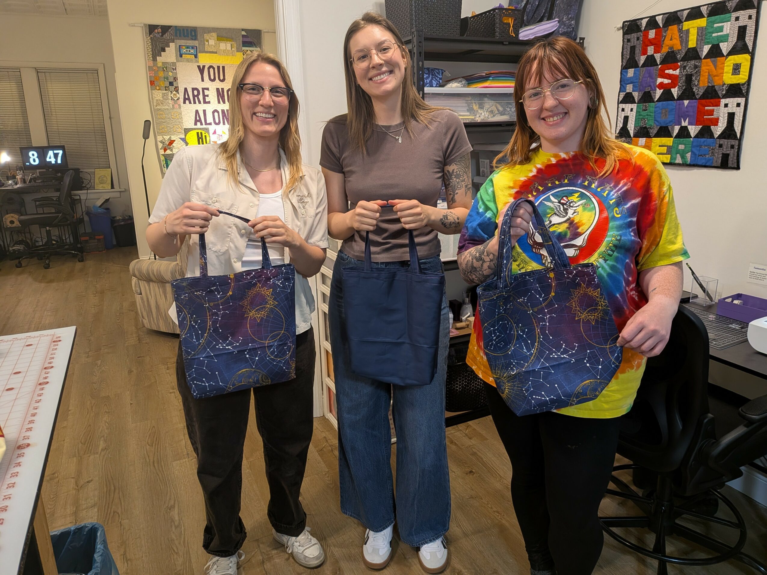 Three women standing indoors, smiling, holding blue bags, with colorful posters on the wall behind them.