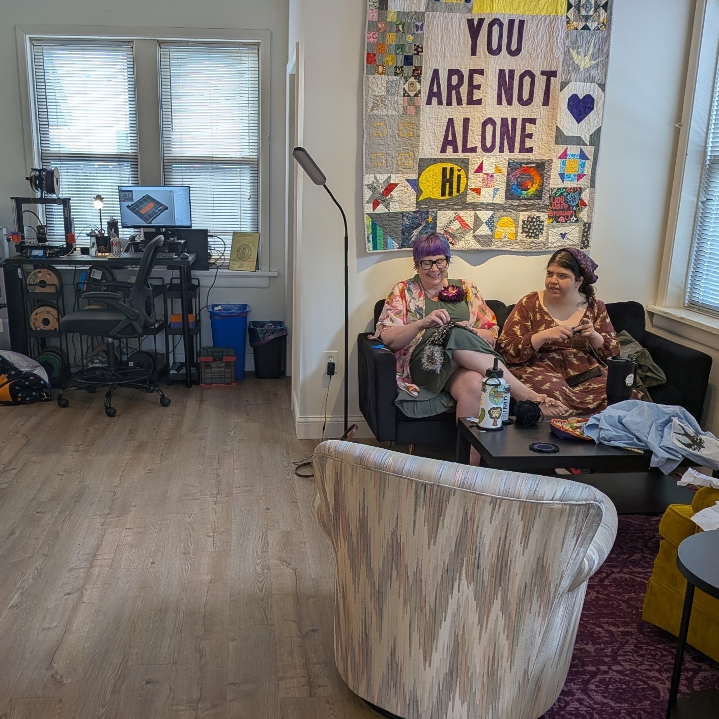 Two women sit on a black couch in a room with a colorful quilted wall hanging that says 'You Are Not Alone.'