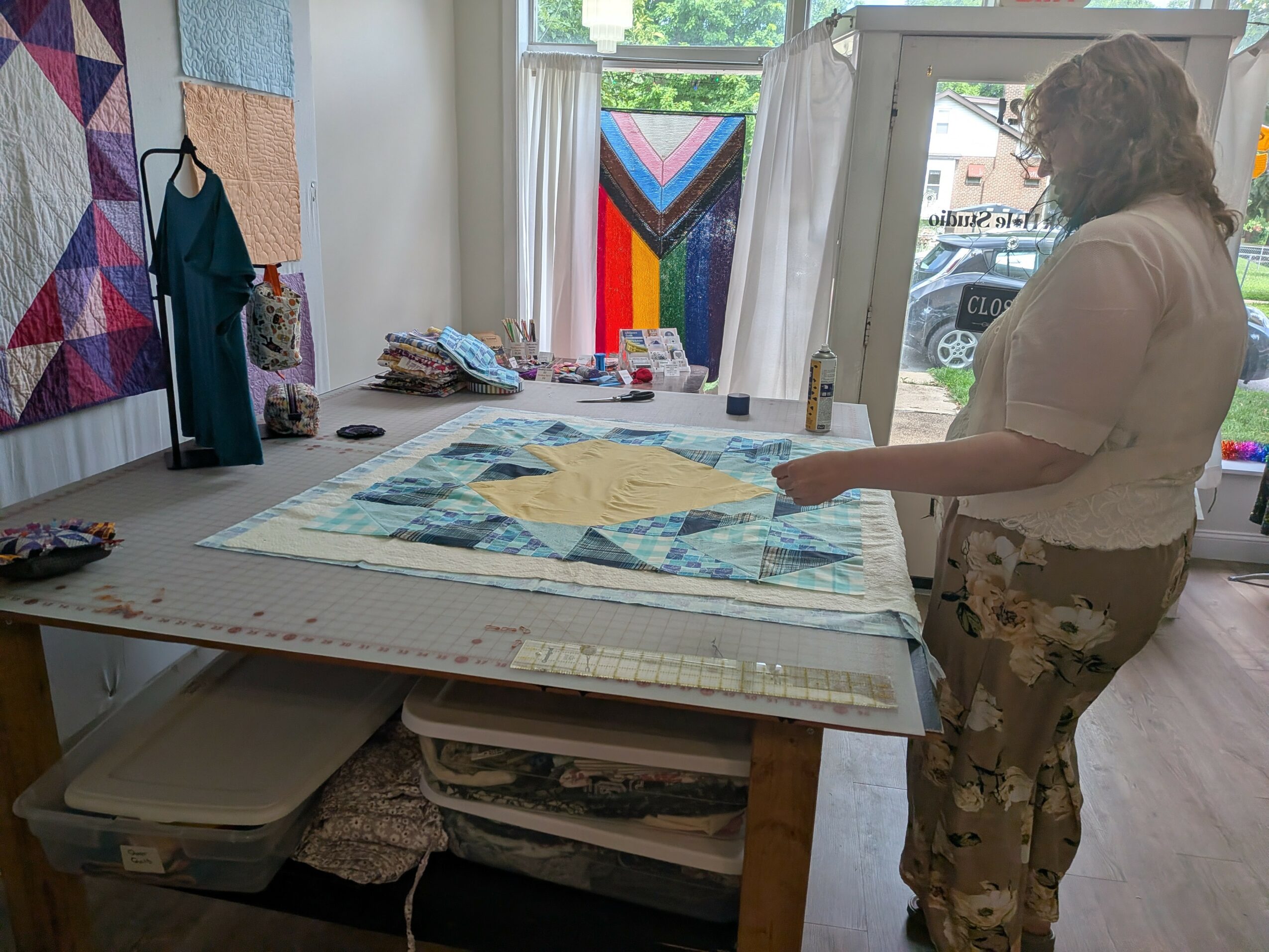 Woman standing at a quilting table with fabric and sewing supplies, window with colorful quilt in background.