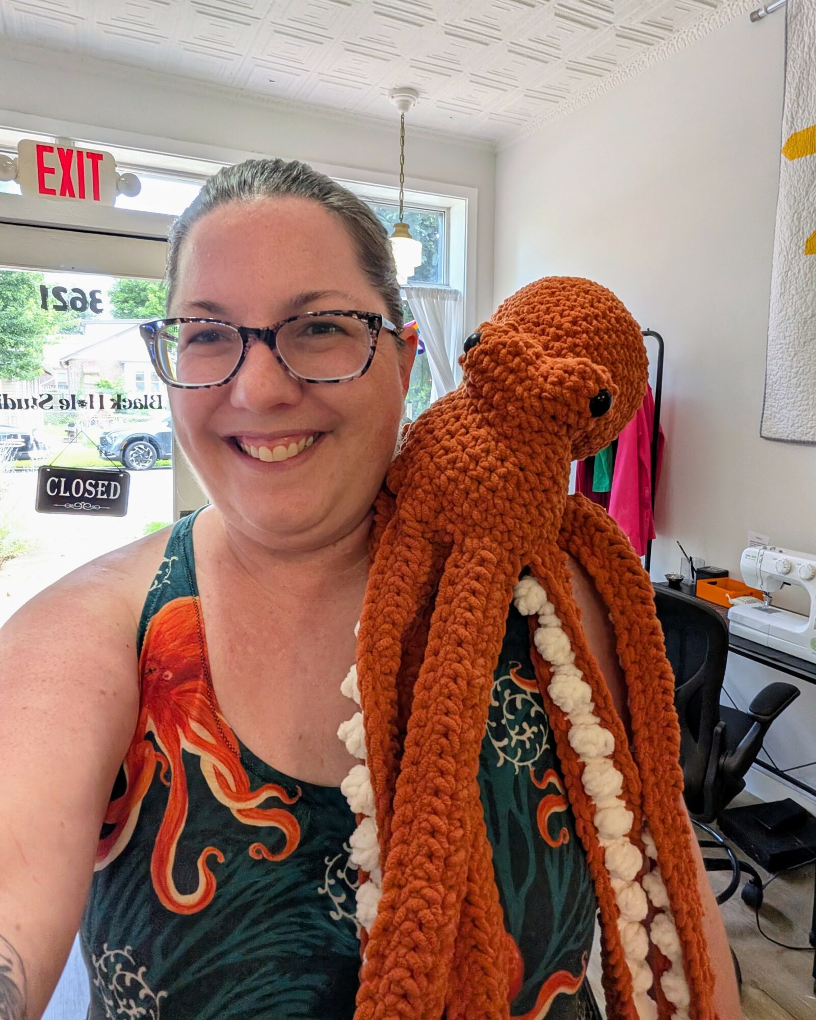 Woman smiling holding a large, orange, crocheted octopus in a room with a window and various objects.