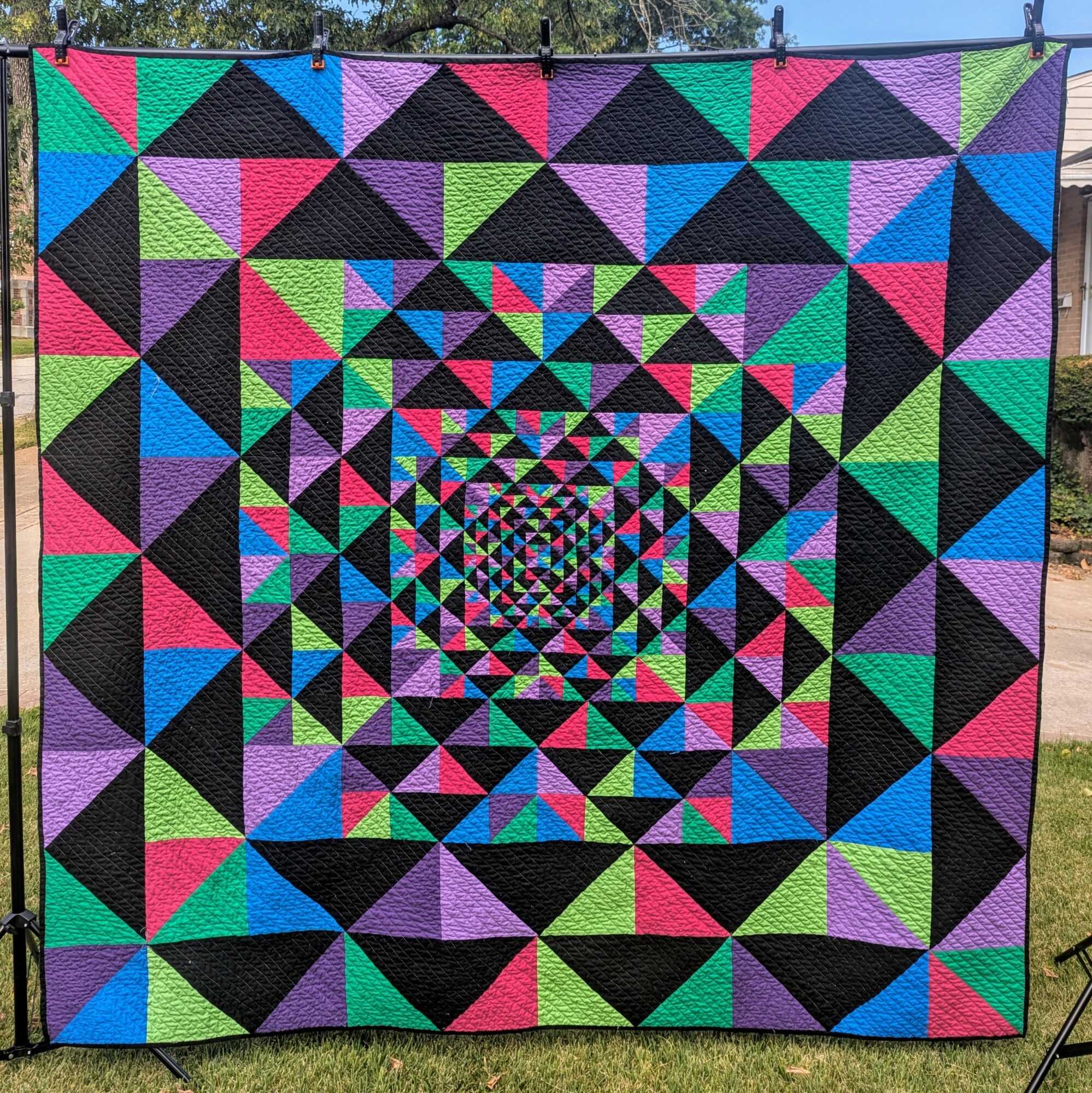 Colorful geometric quilt with concentric triangles and squares, hanging outdoors against a grassy background.