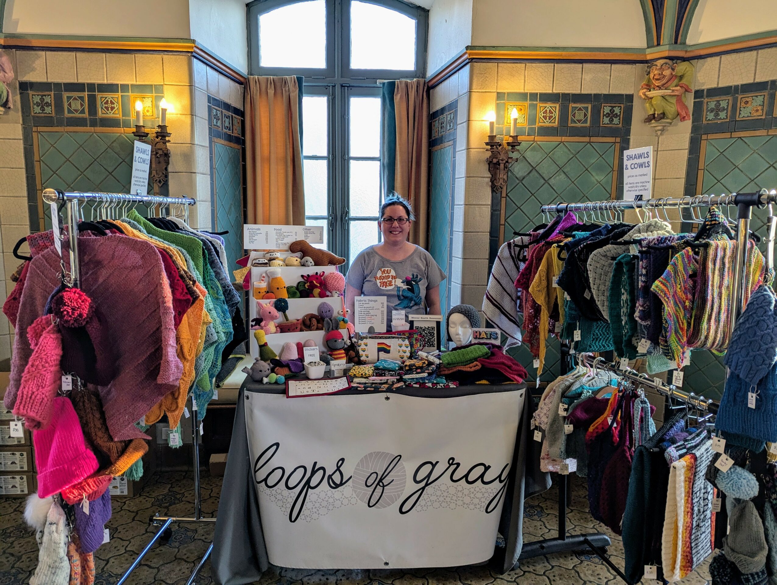 Woman standing behind a table with handmade crafts, surrounded by clothing racks in a decorated room.