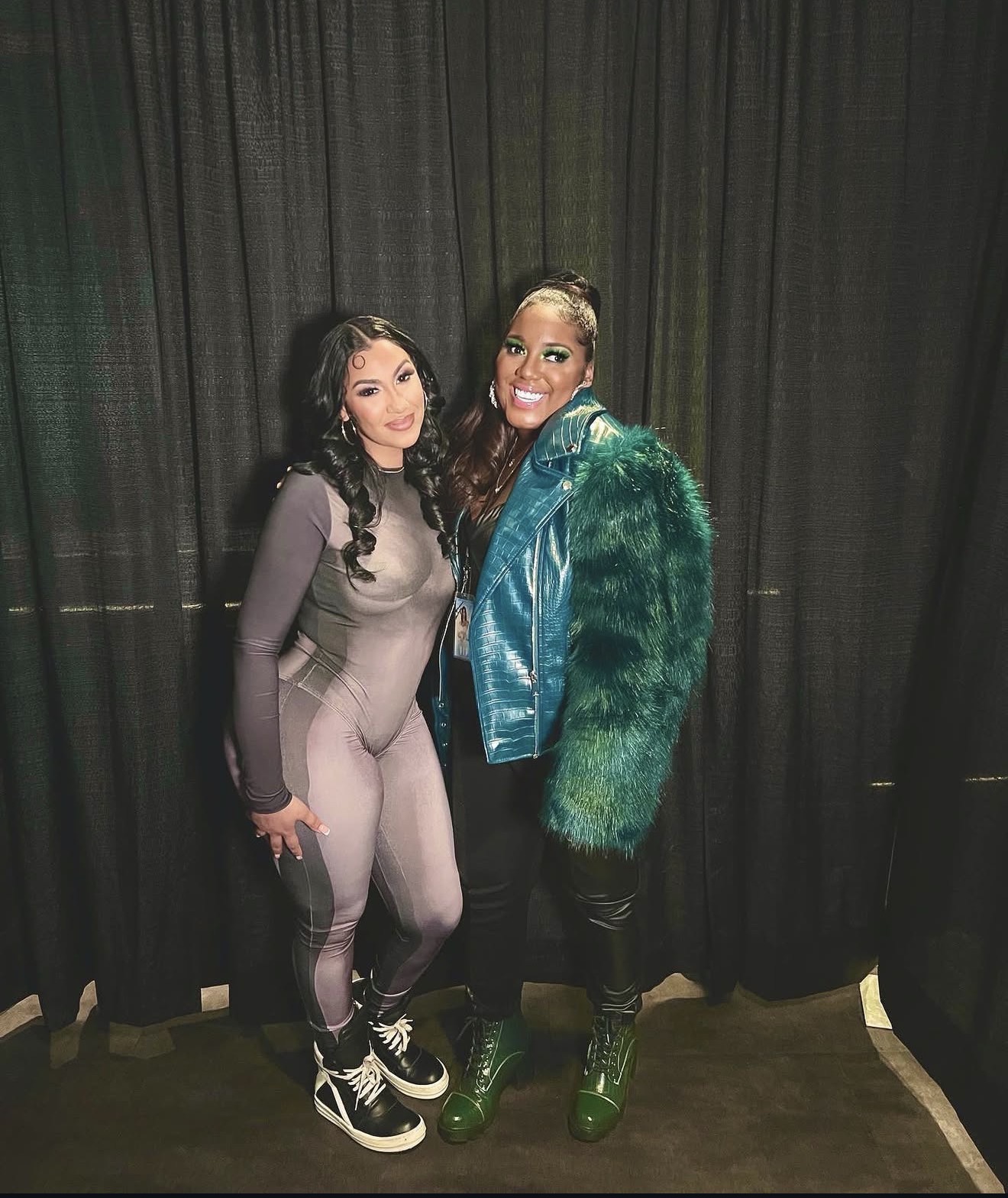 Two women standing together in front of a black curtain, smiling, one in a gray jumpsuit and the other in a green and blue jacket.
