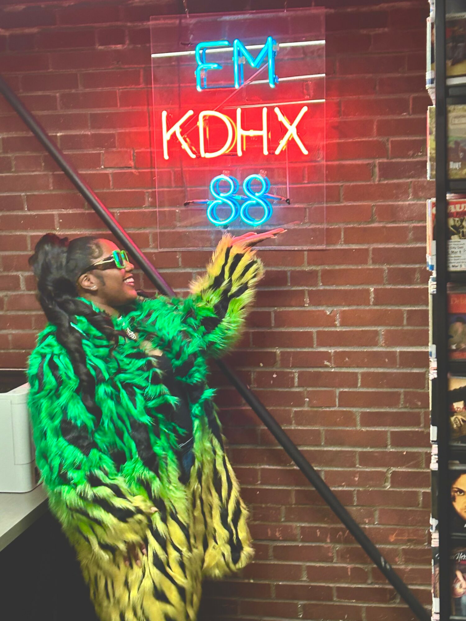 Person wearing a green and yellow patterned coat, pointing at a neon sign on a brick wall, smiling.