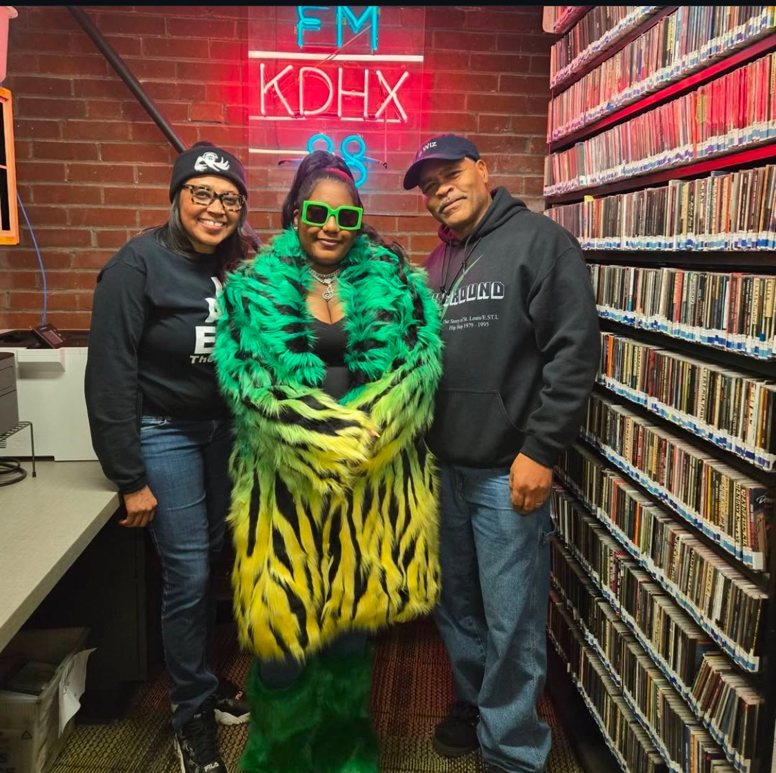 Three people standing in a room with a brick wall and shelves of CDs, one wearing a bright green and yellow tiger-striped coat, all smiling.