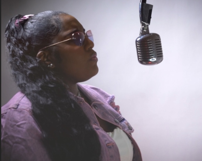 Woman with glasses and braided hair in front of microphone, facing left, against a plain background.