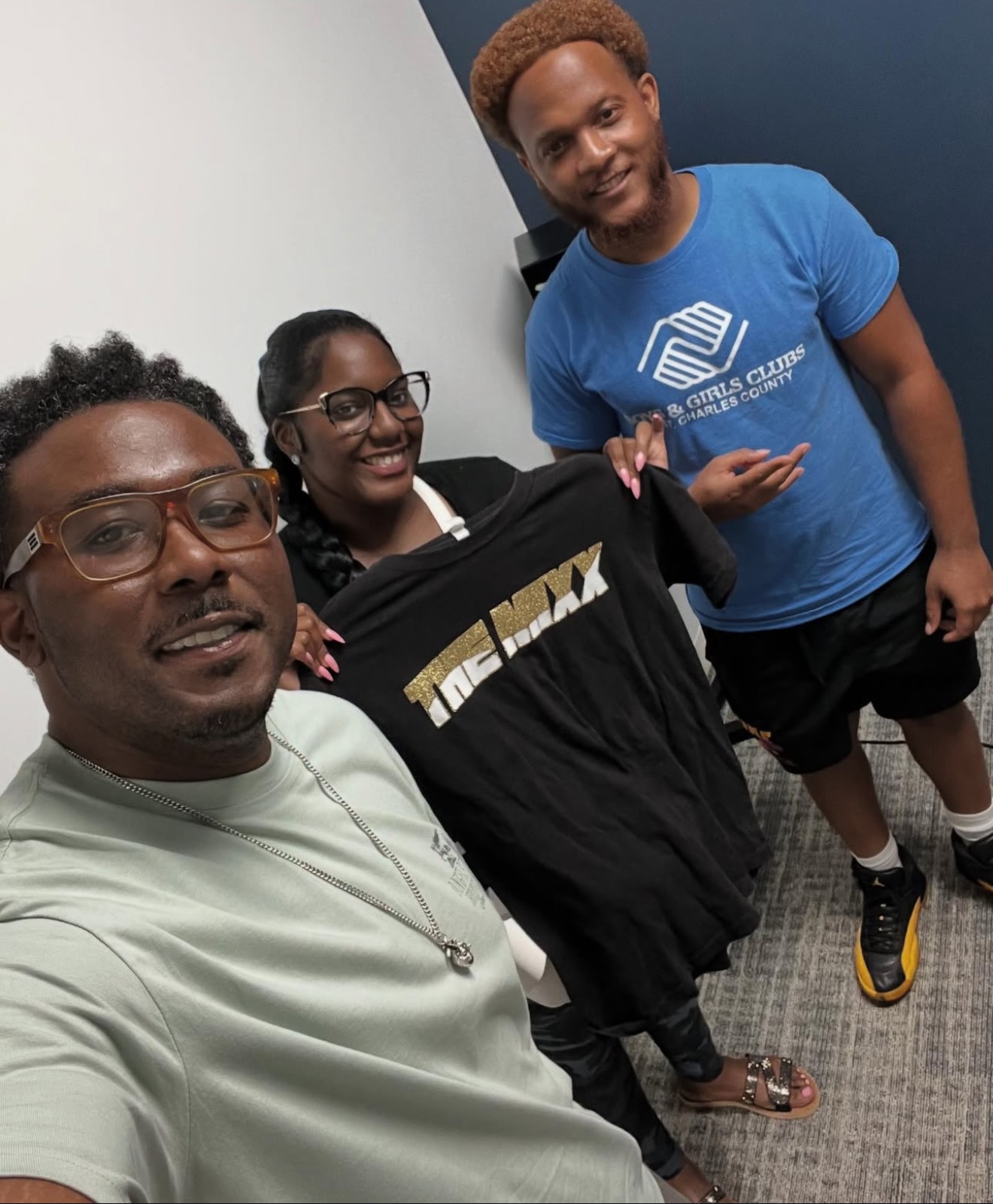 Three people smiling indoors, one holding a black T-shirt with gold text, two wearing glasses, one in a blue T-shirt, all standing close together.