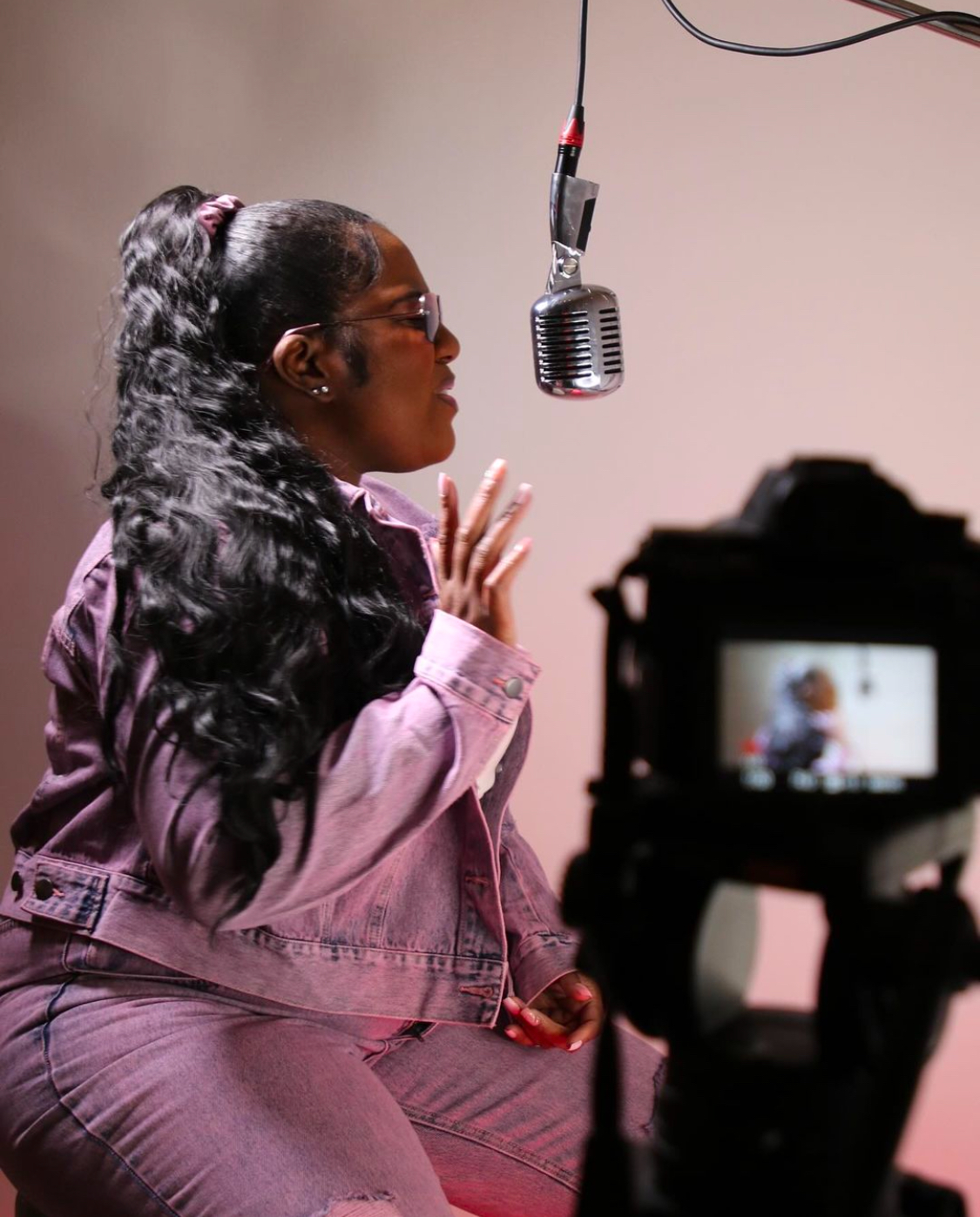 Woman with long curly hair and glasses speaking into a microphone, with a camera recording her, in a studio setting.