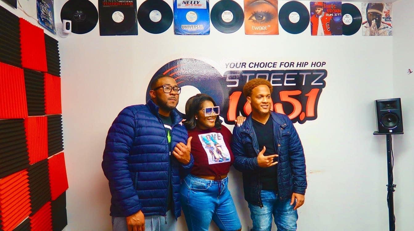 Three people stand in front of a wall with vinyl records and a radio station logo, smiling and posing.