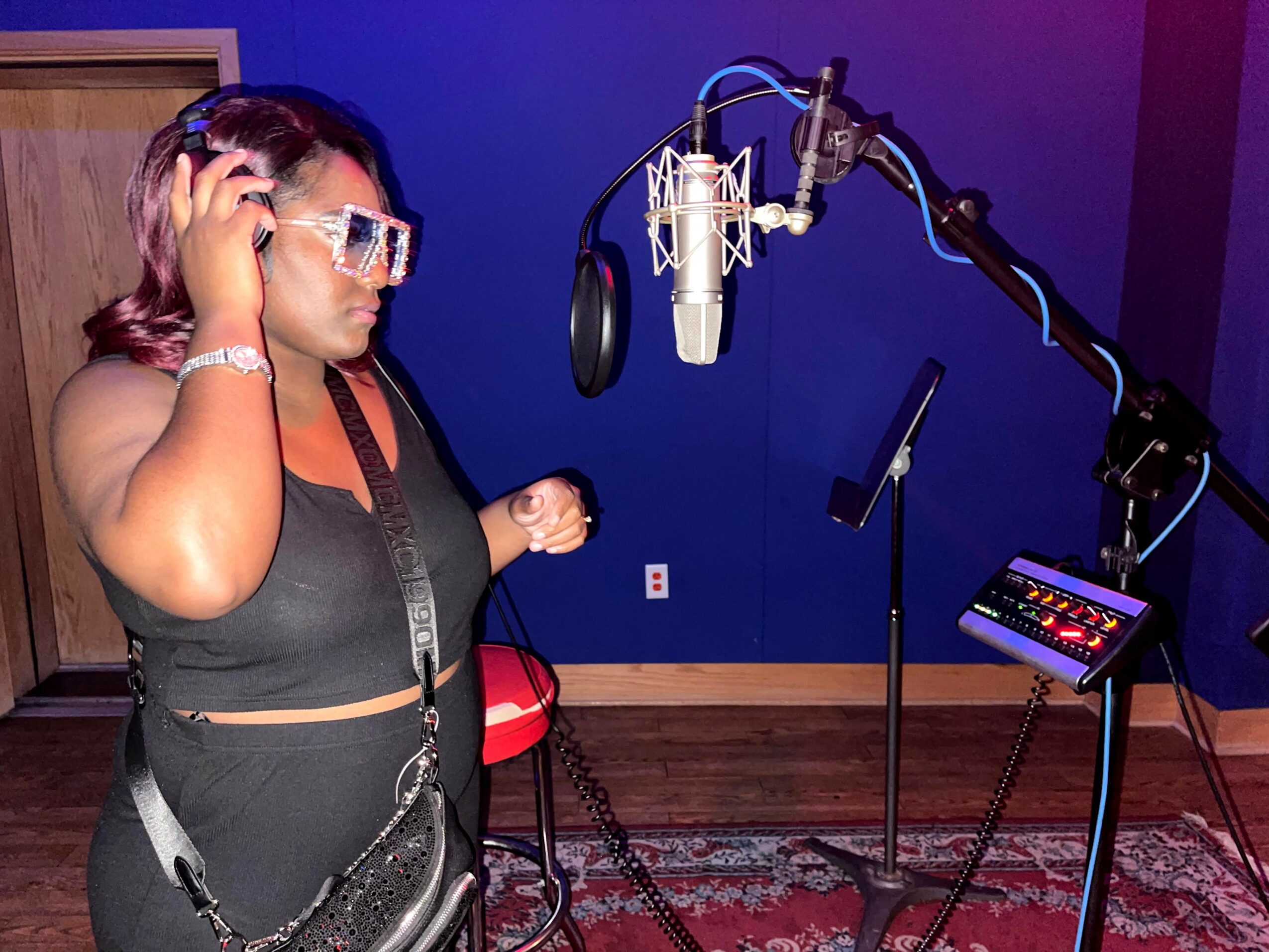 Woman in a recording studio wearing headphones and glasses, standing near a microphone and music equipment.