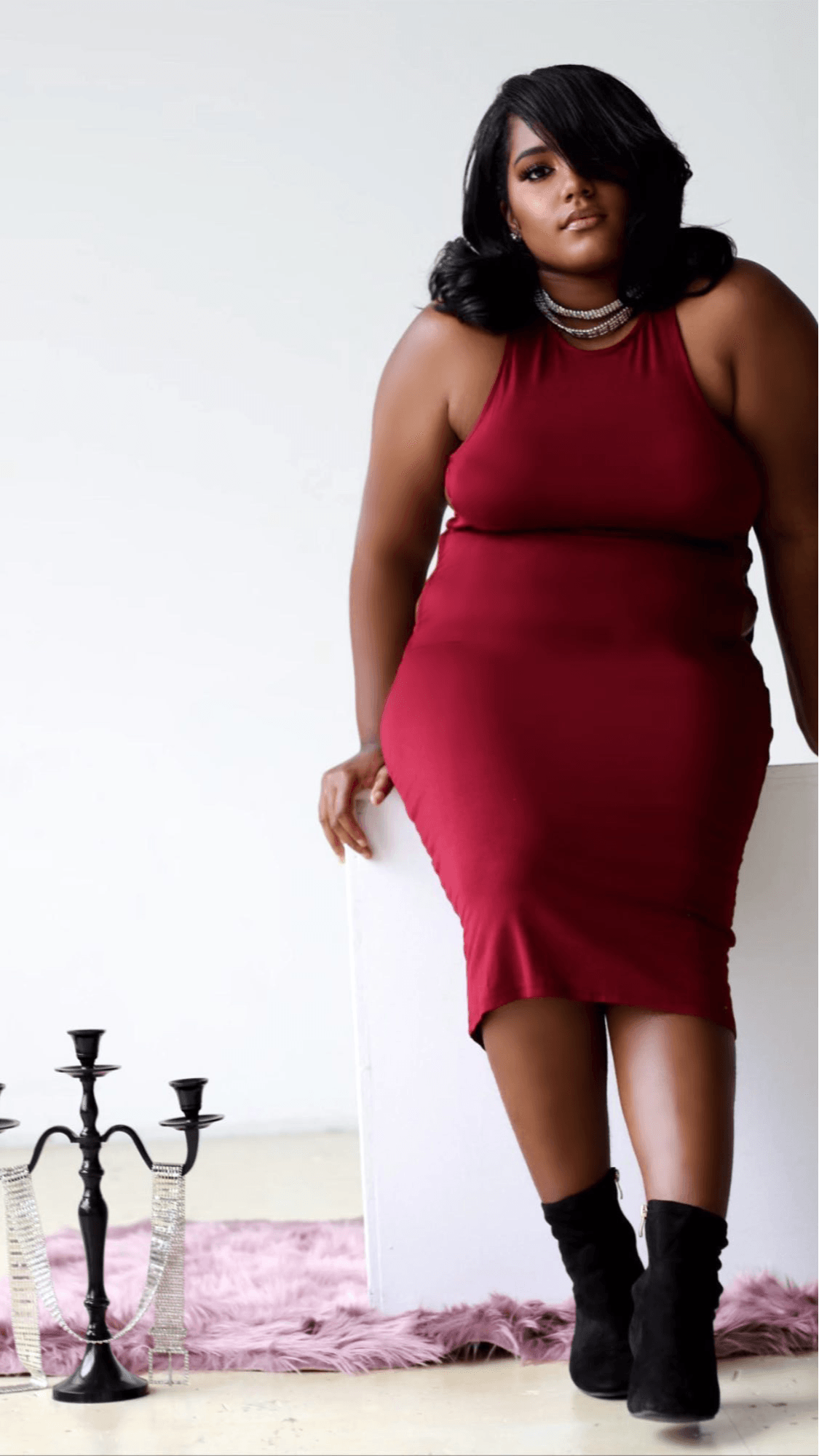 Woman in red dress with black boots standing indoors near a pink rug and a black candelabra.
