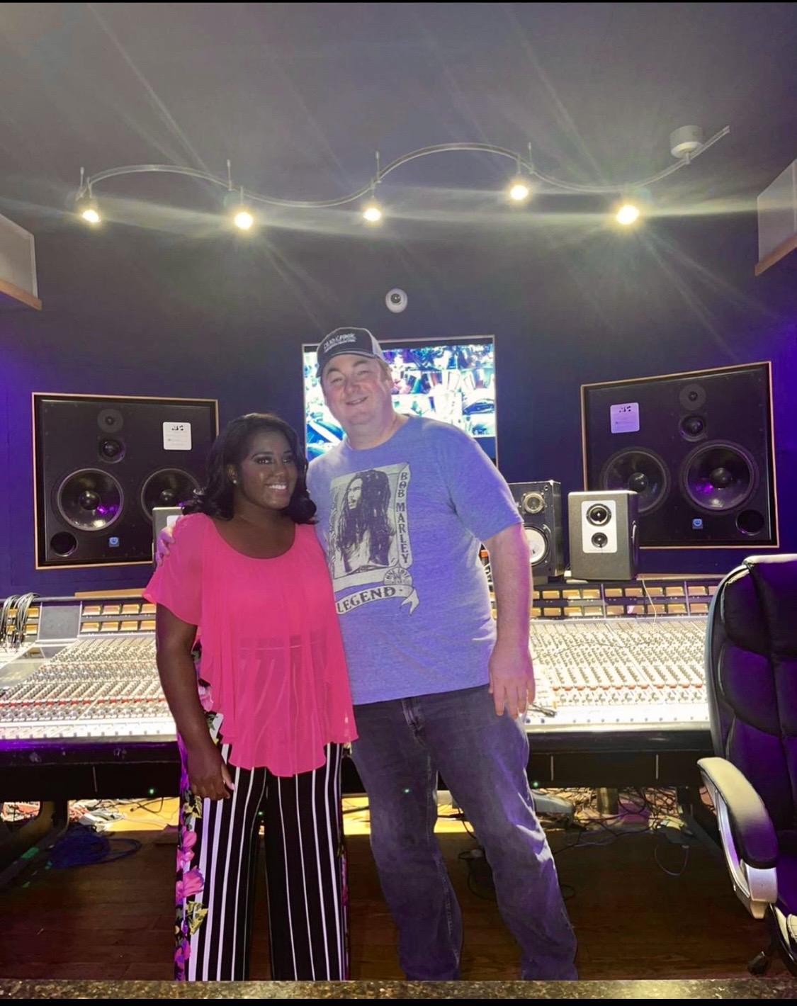 Two people standing in a recording studio with large speakers and mixing console, smiling at the camera.